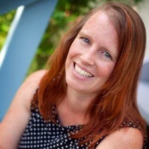 Headshot of a white woman with long red hair smiling and wearing a black and white polka-dotted blouse with no sleeves