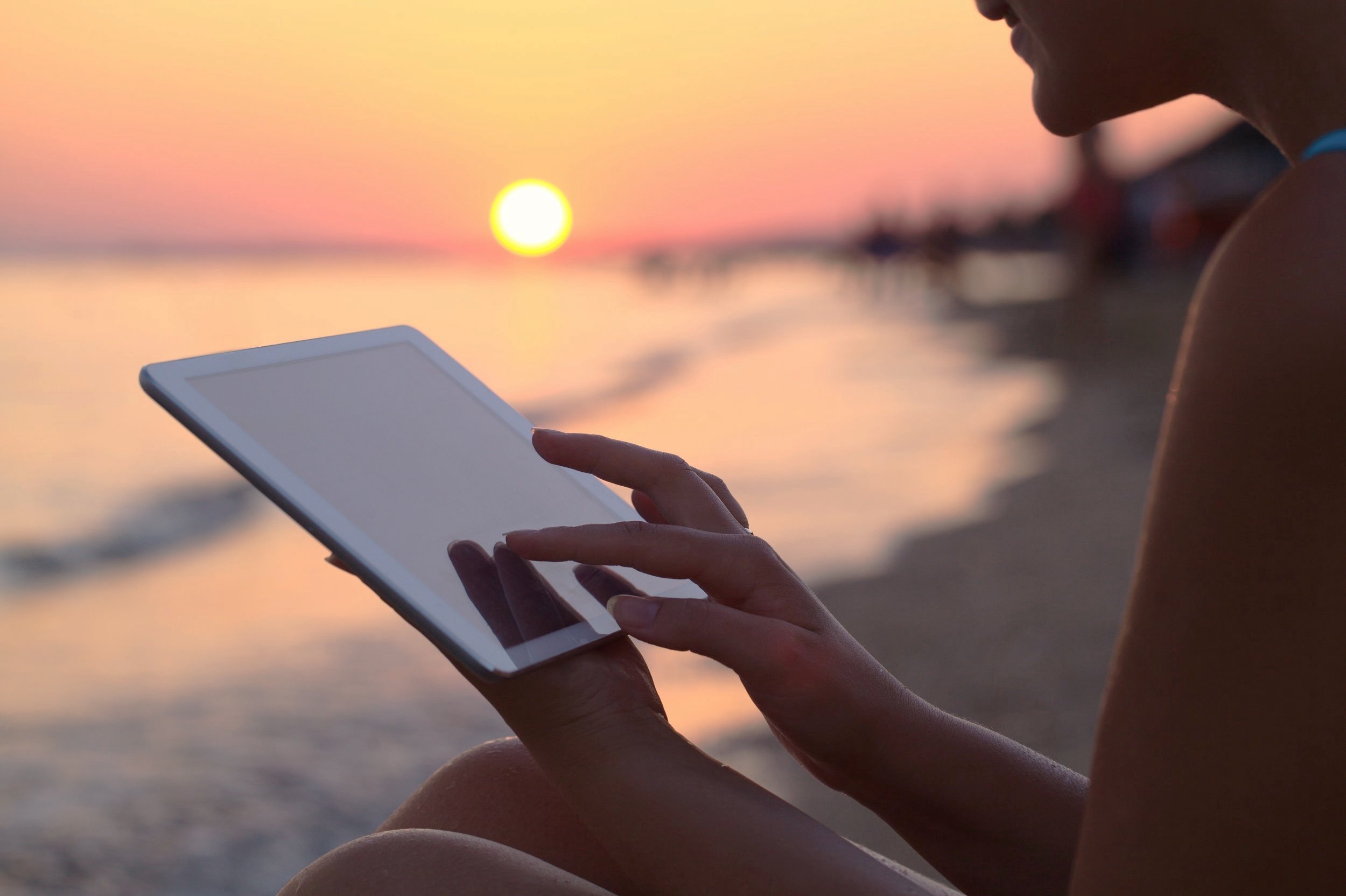 A person sitting on the beach using a tablet during sunset, with the sun near the horizon over the water.