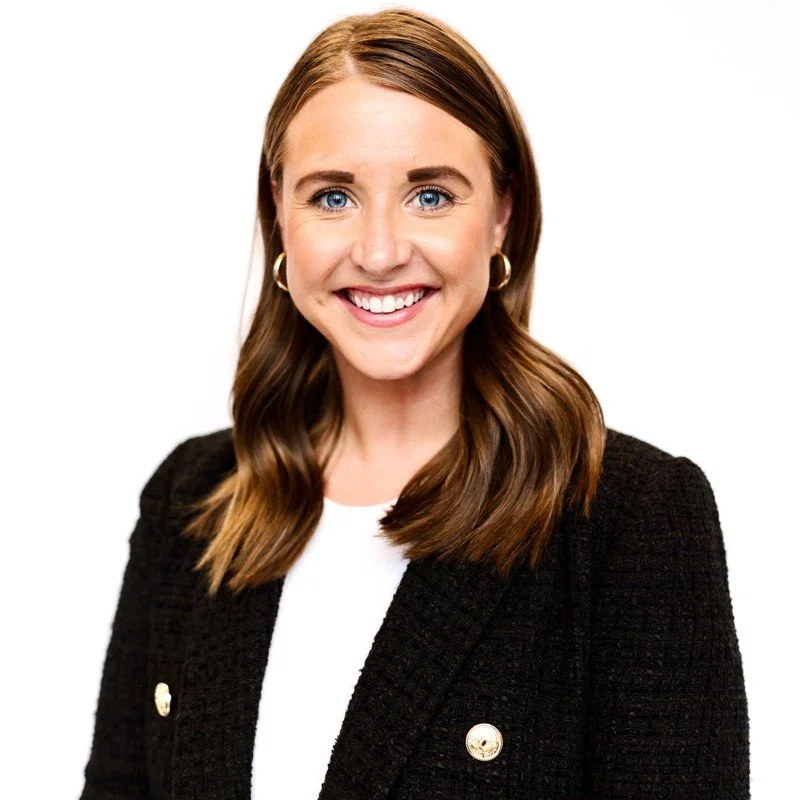 A smiling woman with shoulder-length brown hair, blue eyes, gold hoop earrings, wearing a black blazer over a white top, against a white background.