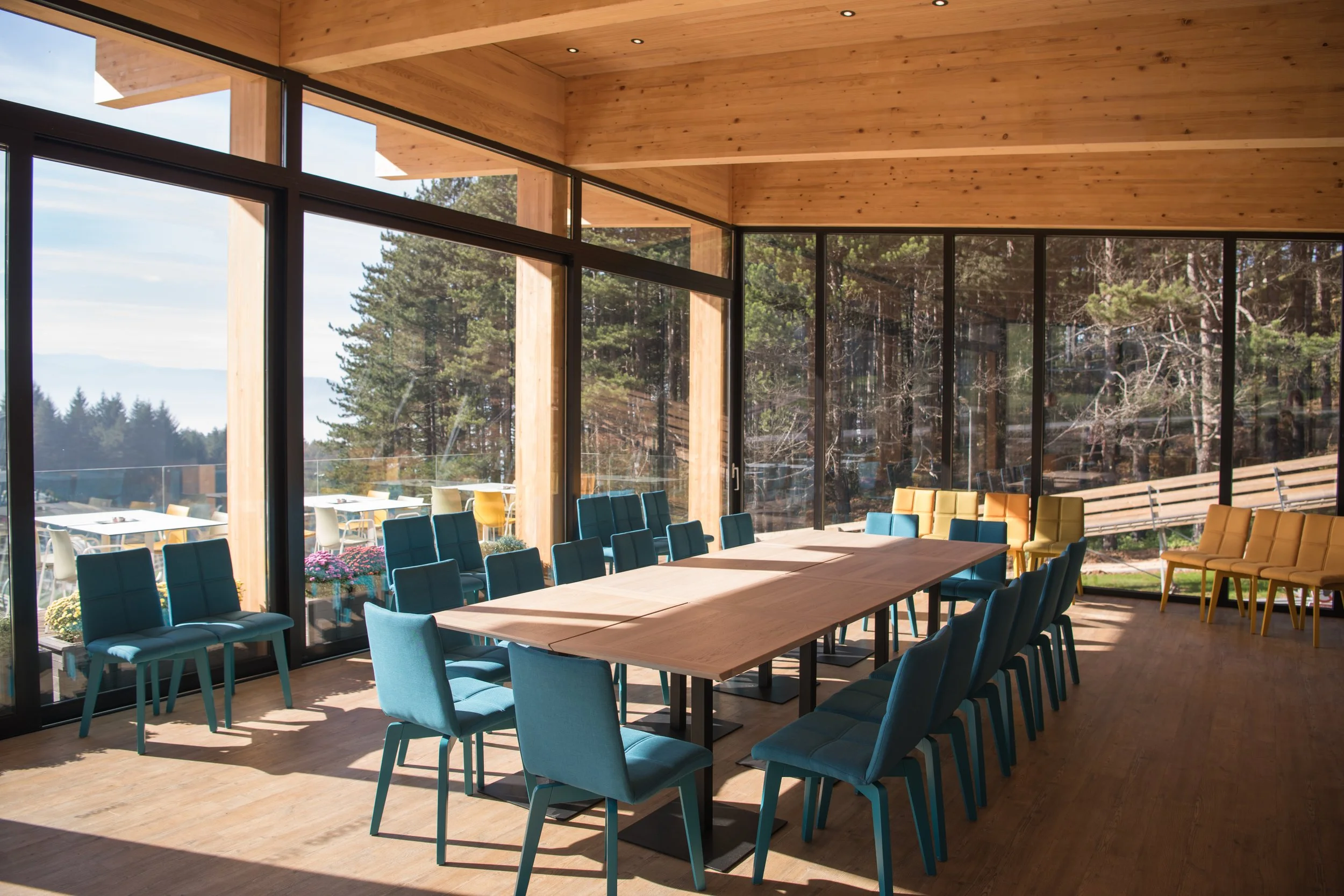 Sunlit conference room with a long wooden table surrounded by blue and yellow chairs, large glass windows showing trees, mountains, and outdoor seating area.