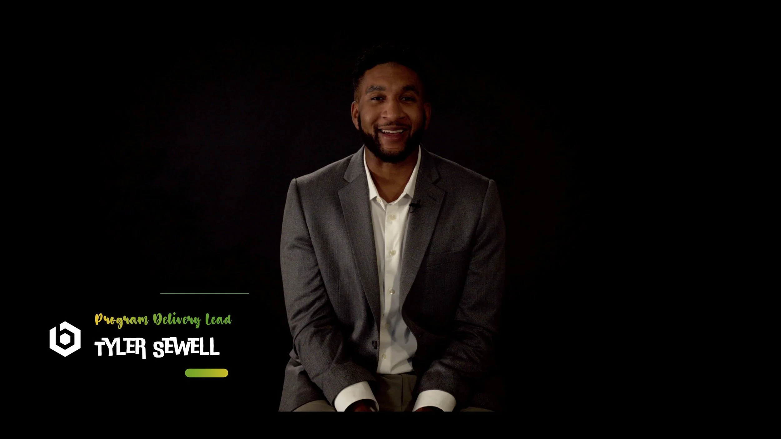 A man in a gray suit and white shirt sitting against a black background, smiling at the camera, with text on the lower left that reads 'Program Delivery Lead TYLER SEWELL'.