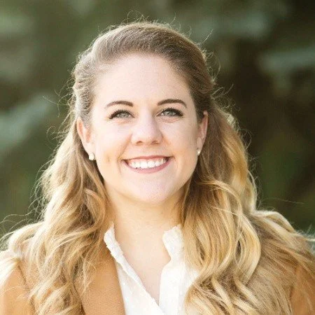 A young woman with long blond hair smiling outdoors, wearing a brown jacket and white shirt.