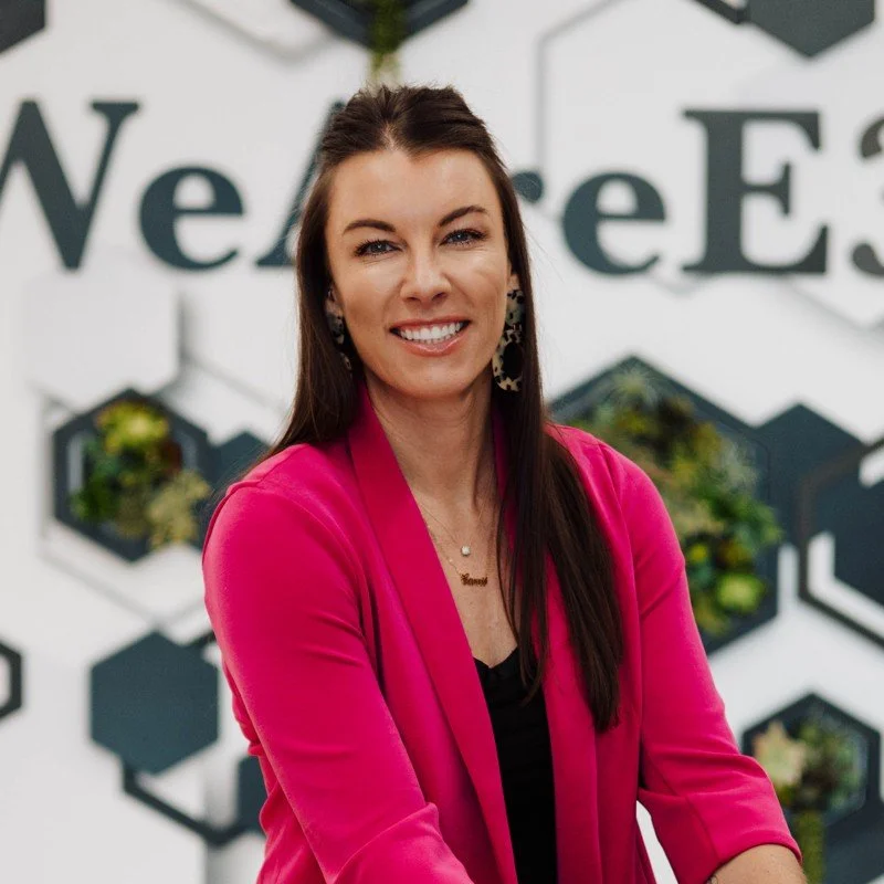 A woman with long dark hair, wearing a bright pink blazer and earrings, smiling in front of a wall with black and white hexagonal wall planters and large letters in the background.