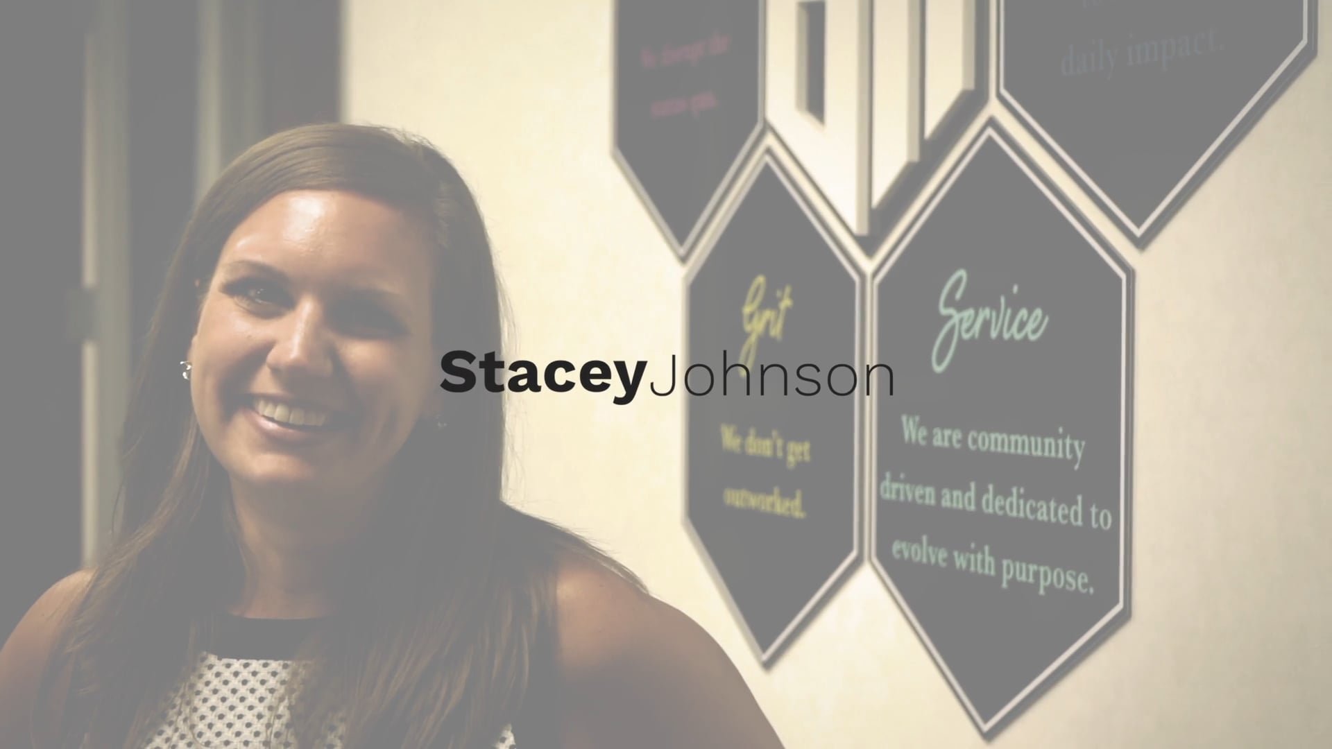 A woman with long brown hair and a white sleeveless top smiling in an office environment with wall decor containing text about community service and mission statements.
