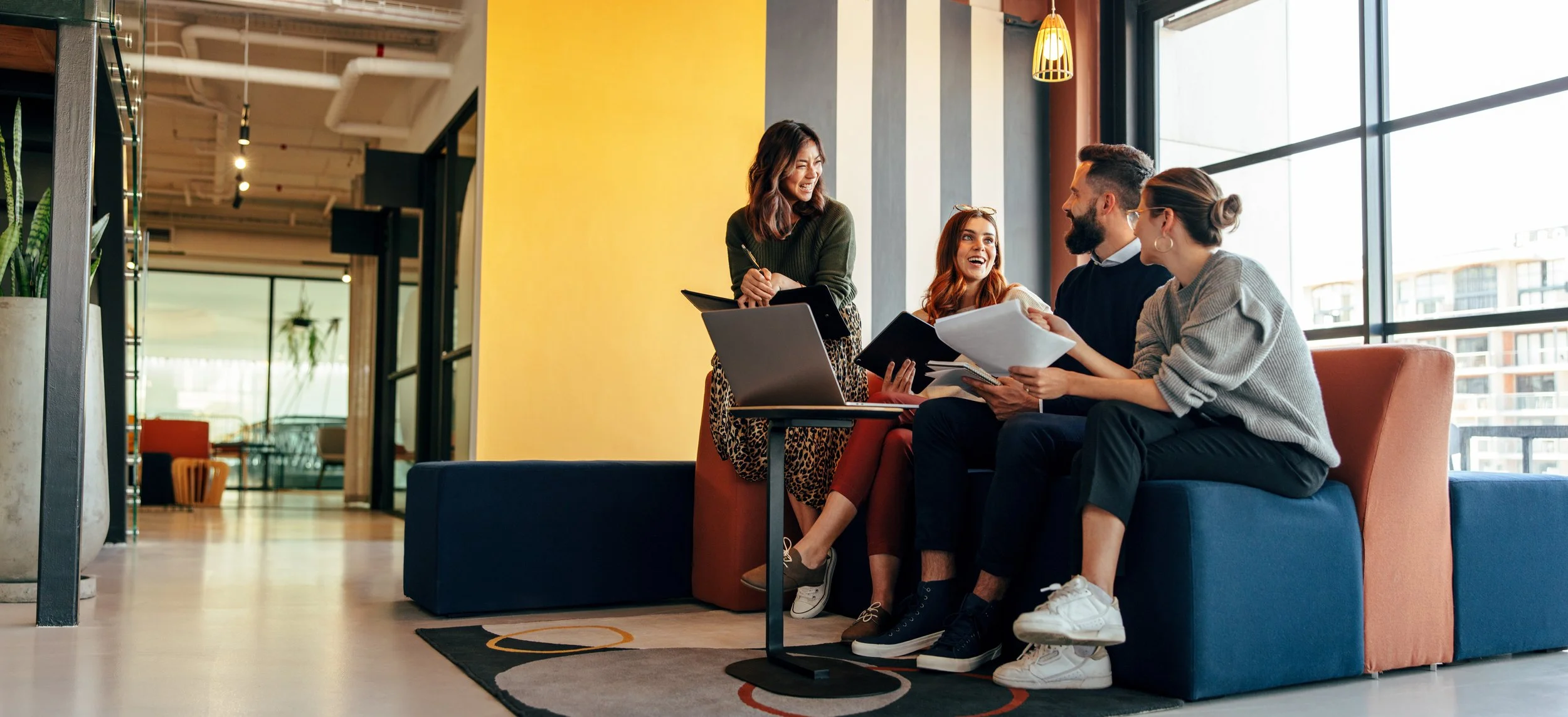 Four diverse young professionals having a discussion in a bright, modern office lounge with large windows and colorful furniture.