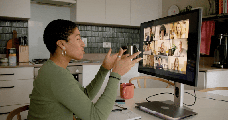 A woman with short curly hair and earrings participates in a video call on a desktop computer in a kitchen. She gestures with her hands, and the screen shows multiple people in the video conference.