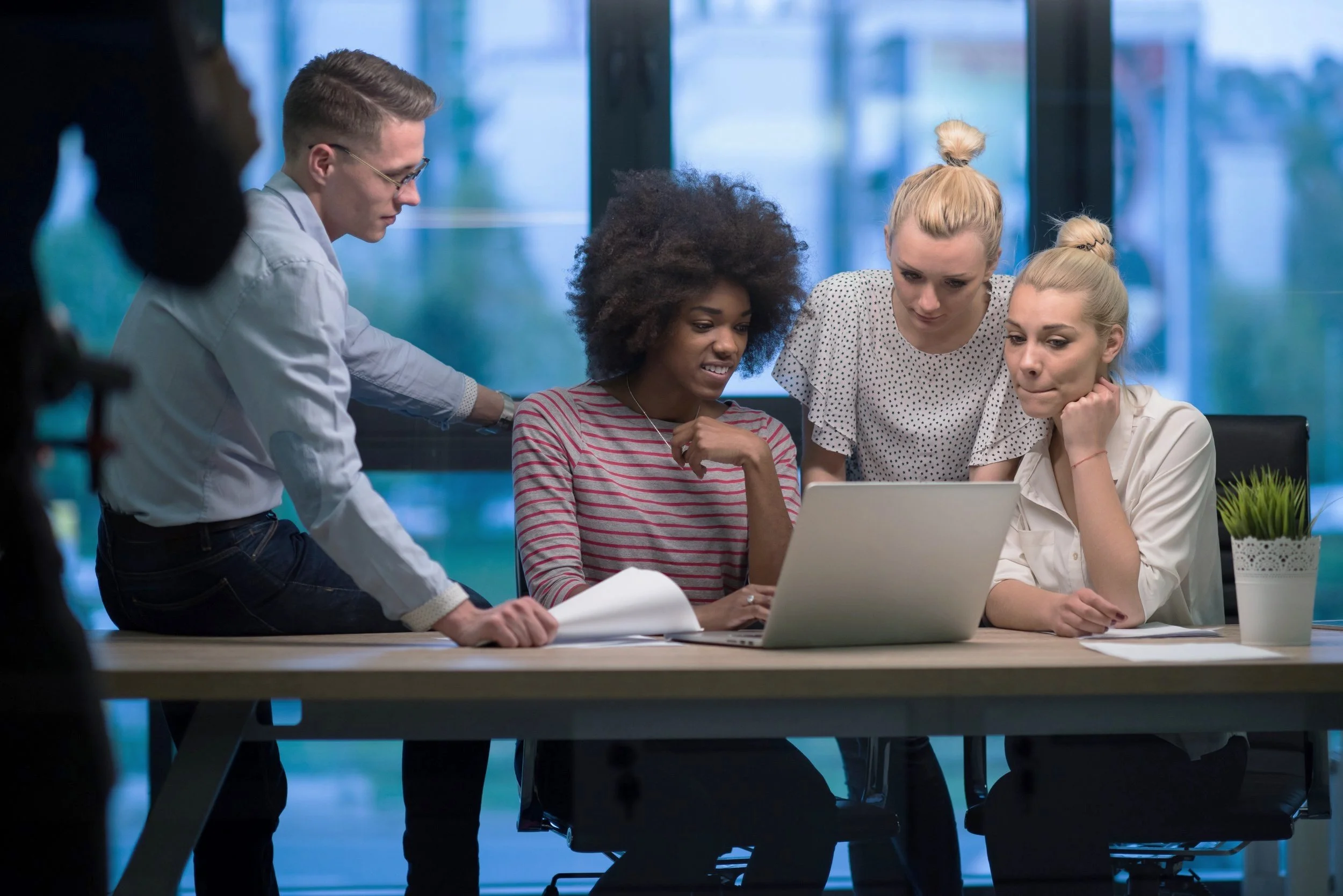 Four people gathered around a laptop at a meeting in a modern office with large windows.