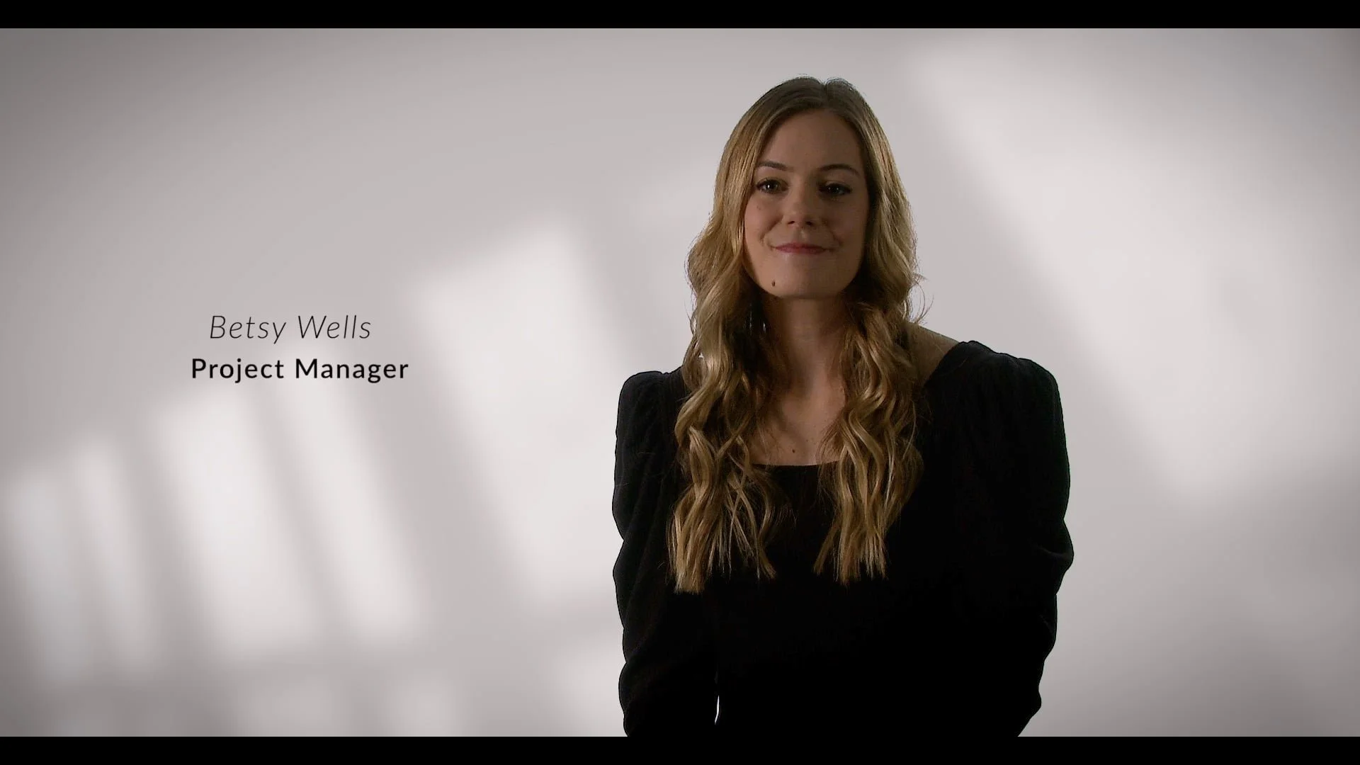 A woman with long wavy blonde hair, wearing a black top, smiling against a plain background, with text identifying her as Betsy Wells, Project Manager.