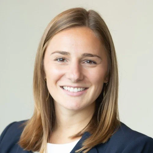 A woman with shoulder-length brown hair, smiling, wearing a dark blazer and a white top, standing against a plain light background.