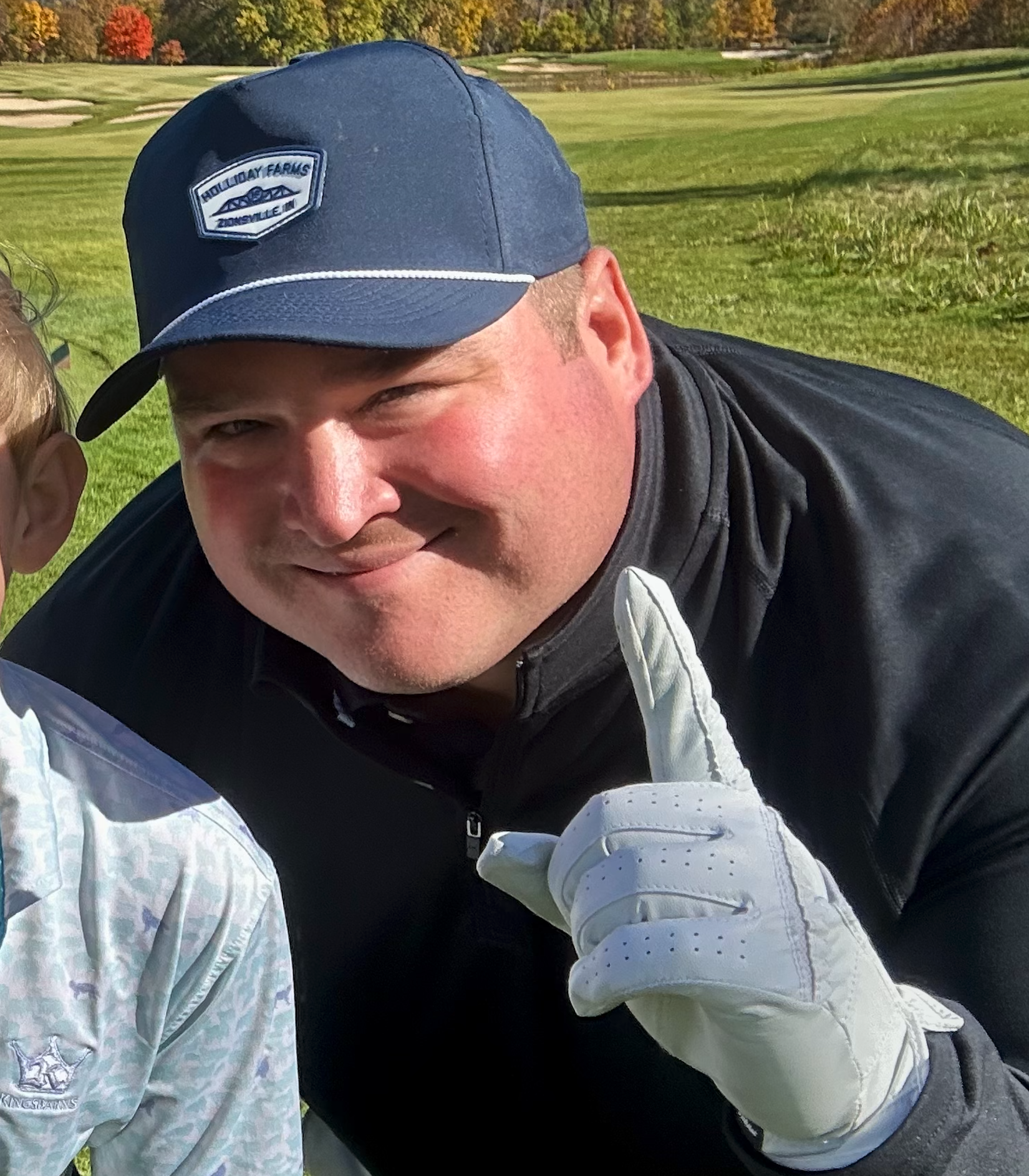 A man in a blue cap and black jacket smiling outdoors on a golf course, holding up one finger.