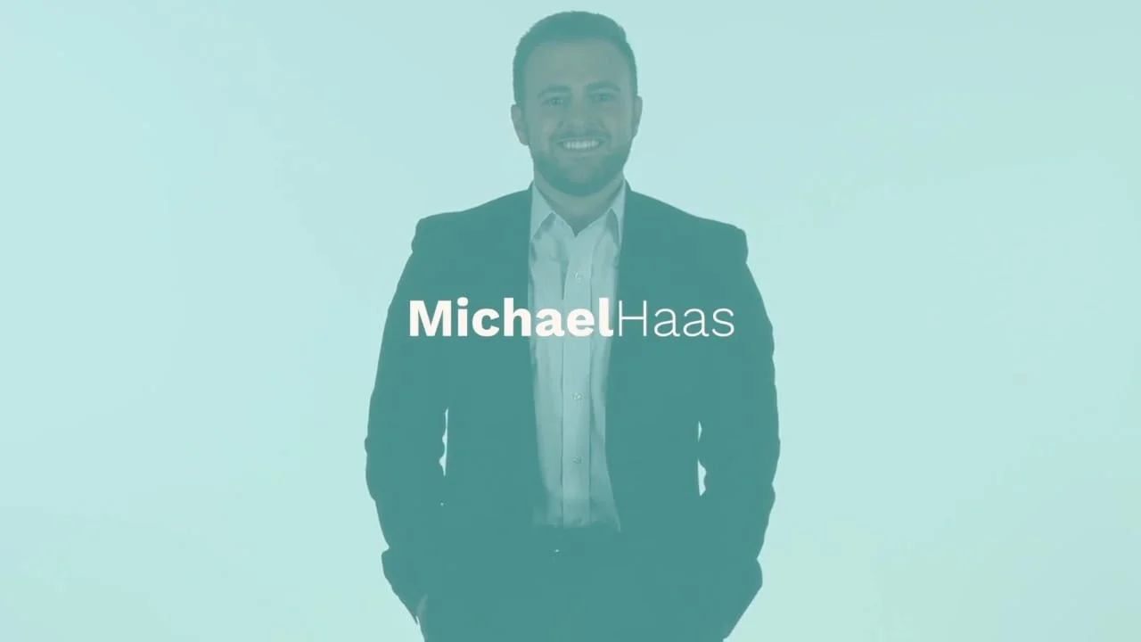 Professional portrait of Michael Haas wearing a suit and smiling against a light blue background.