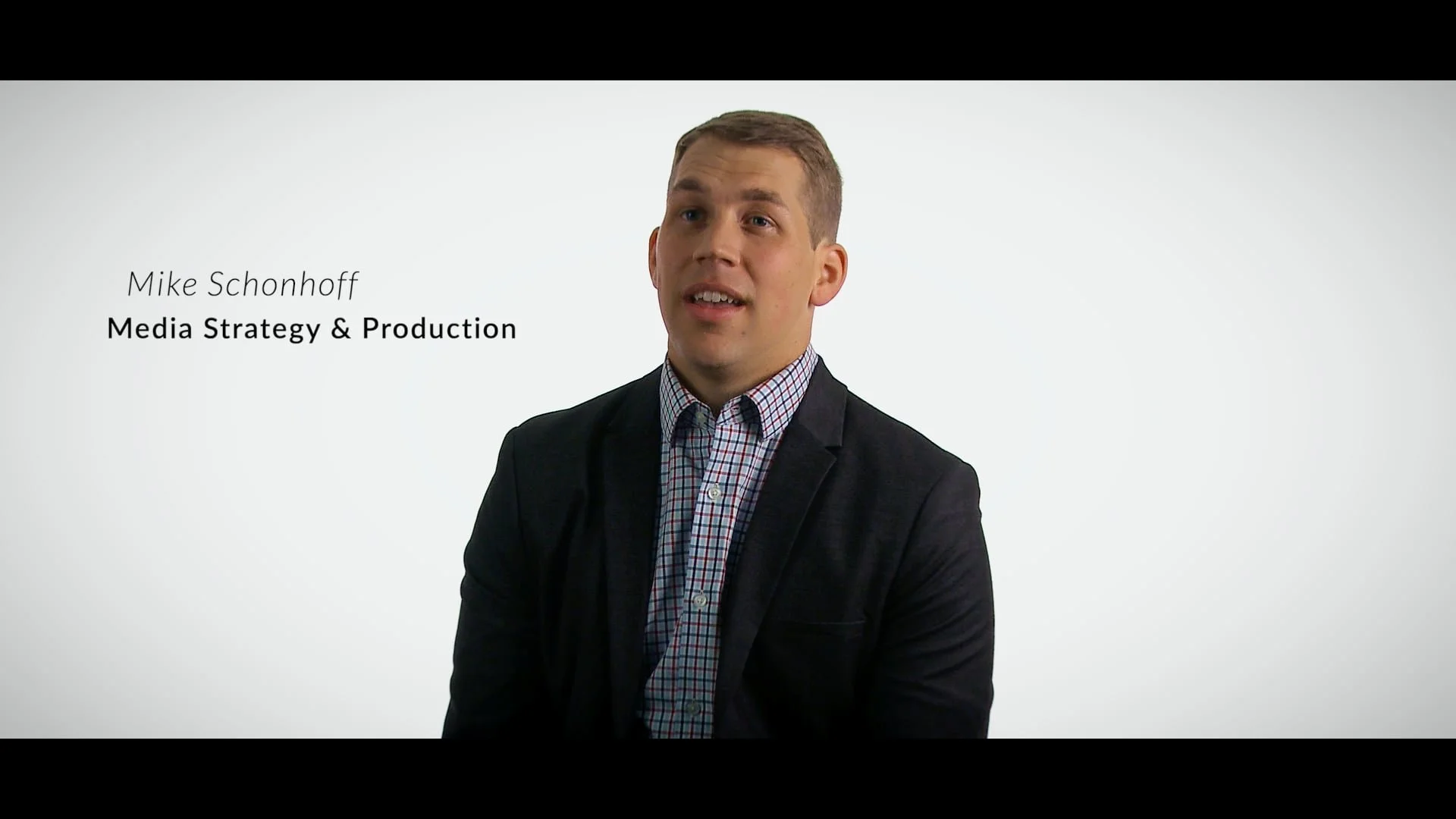 A man in a dark blazer and checkered shirt speaking, with text on the left that reads 'Mike Schonhoff, Media Strategy & Production' against a plain white background.