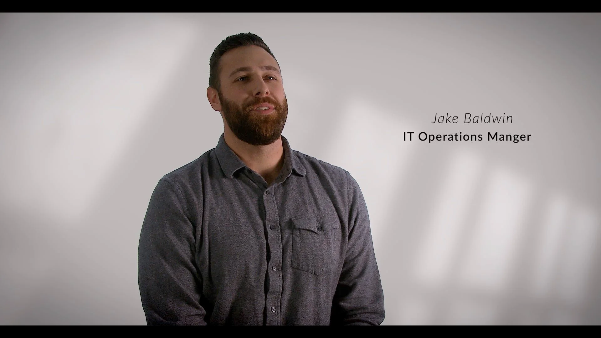 A man with a beard, wearing a gray shirt, standing against a light gray background with text that reads 'Jake Baldwin, IT Operations Manager'.