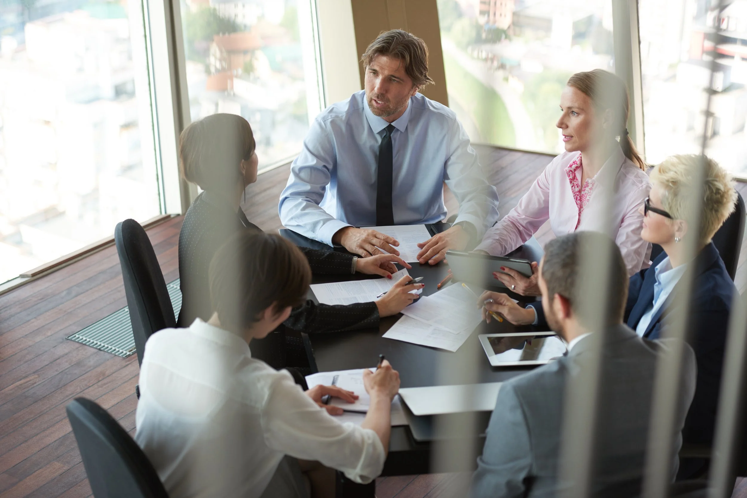 Business meeting in a conference room with six professionals sitting around a table, discussing, with large windows overlooking a cityscape in the background.