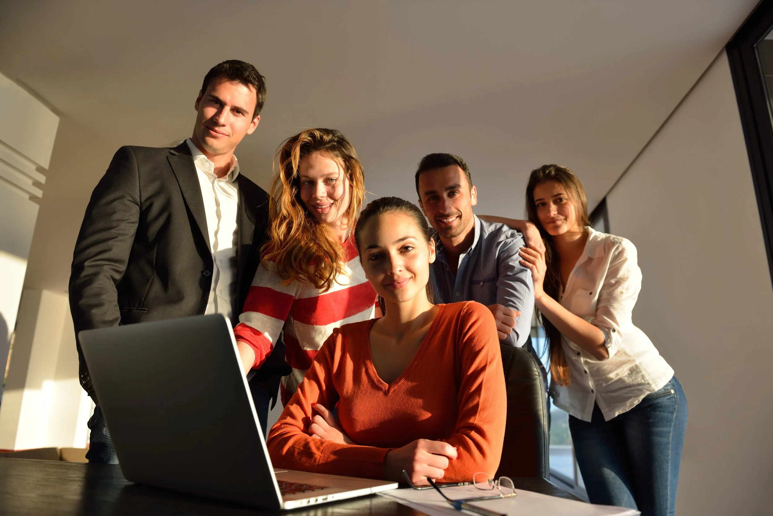 Group of five young professionals gathered around a laptop in a modern office.