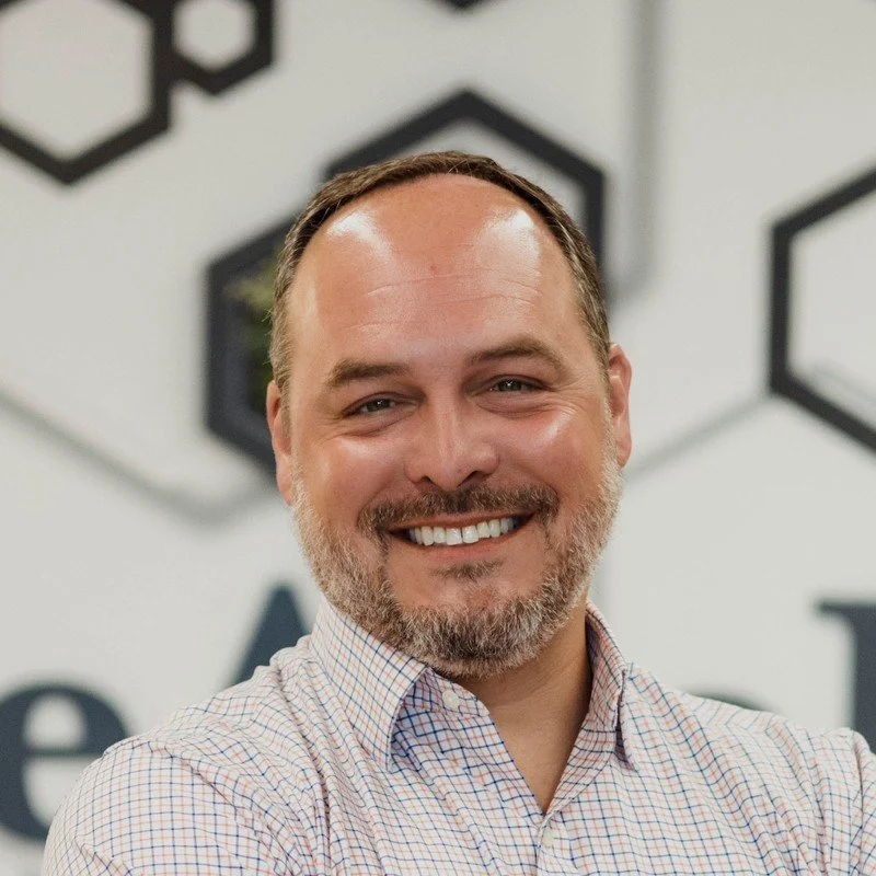 A smiling man with a beard and short brown hair, wearing a checkered button-up shirt, standing in front of a wall with black and white geometric patterns.
