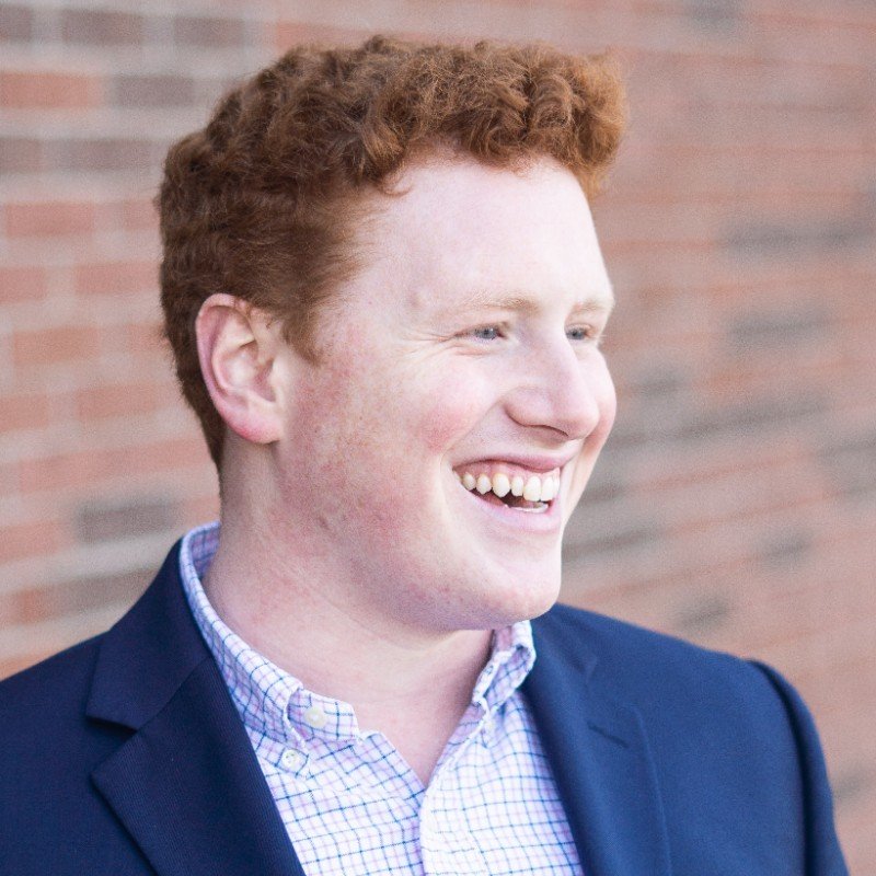 Smiling man with red hair wearing a navy blazer and a checkered shirt, standing outdoors against a brick wall.