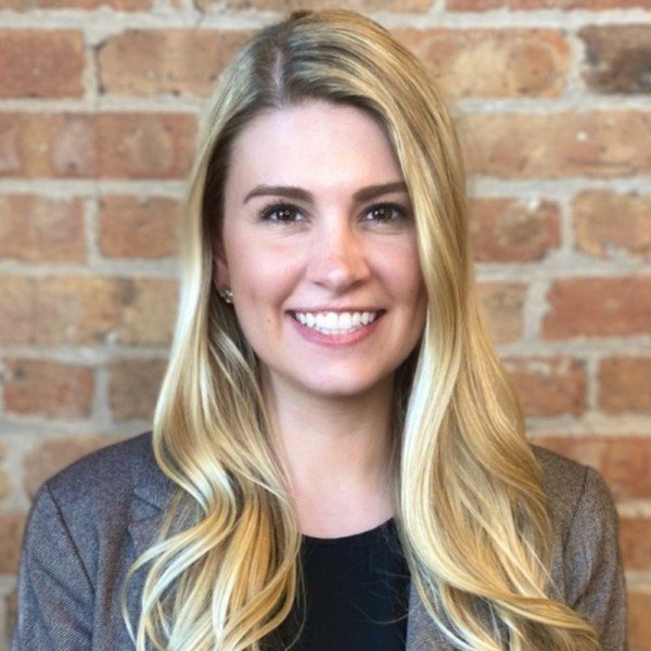 Young woman with long blonde hair smiling, wearing a gray blazer and black top, standing in front of a brick wall.