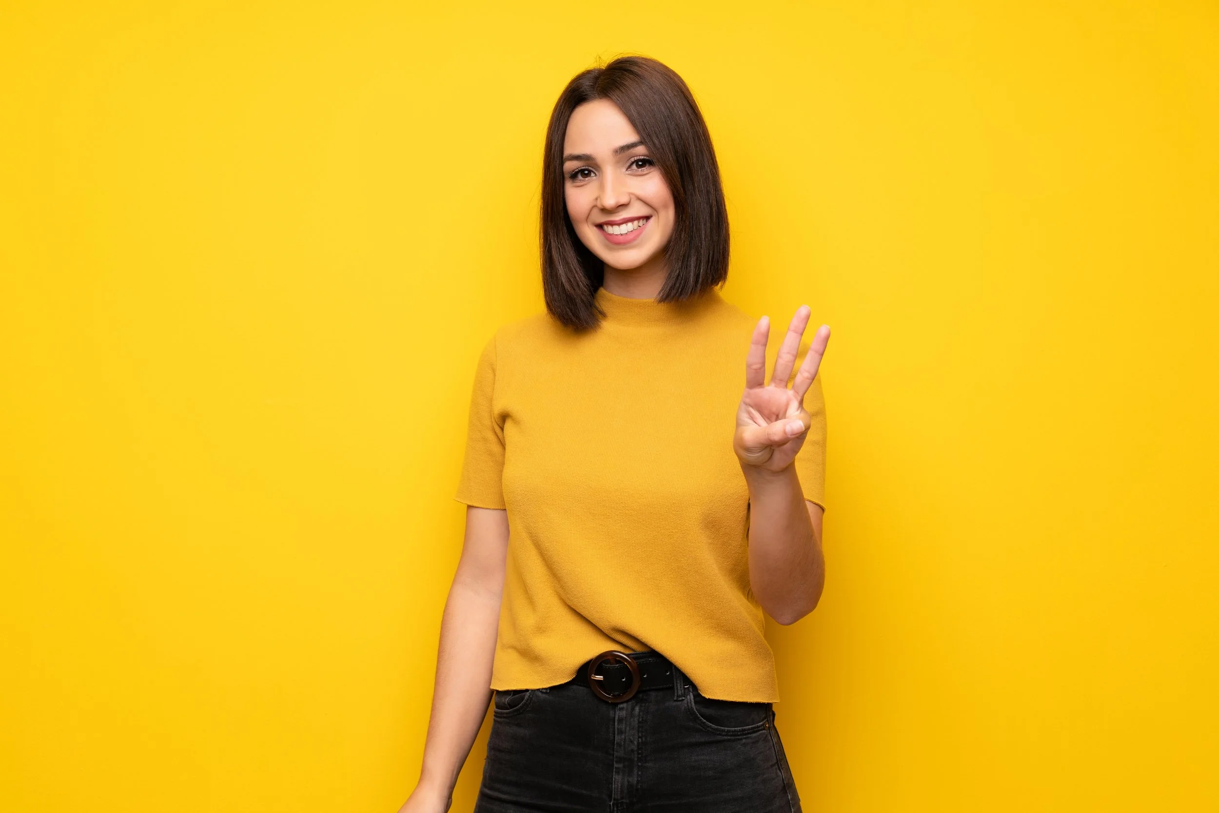 A smiling young woman with shoulder-length brown hair wearing a yellow t-shirt and black jeans stands against a yellow background, holding up three fingers.