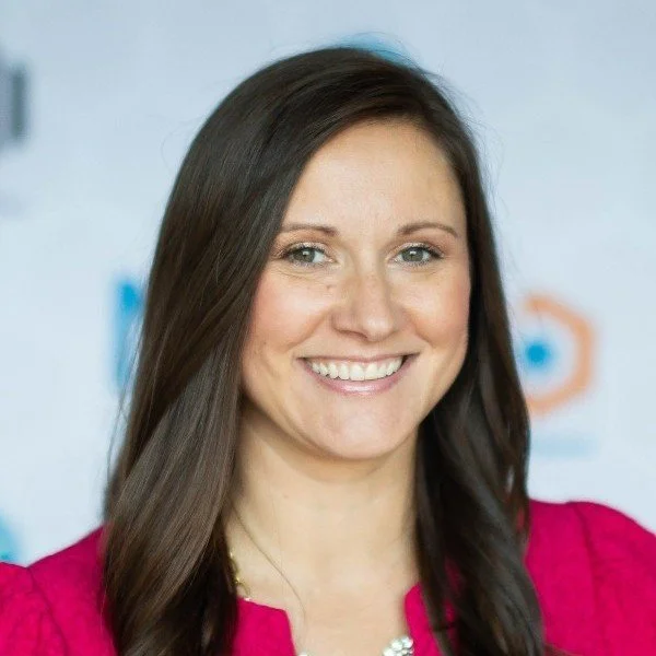 A woman with long brown hair smiling, wearing a pink top, and standing in front of a blurred background with logos.