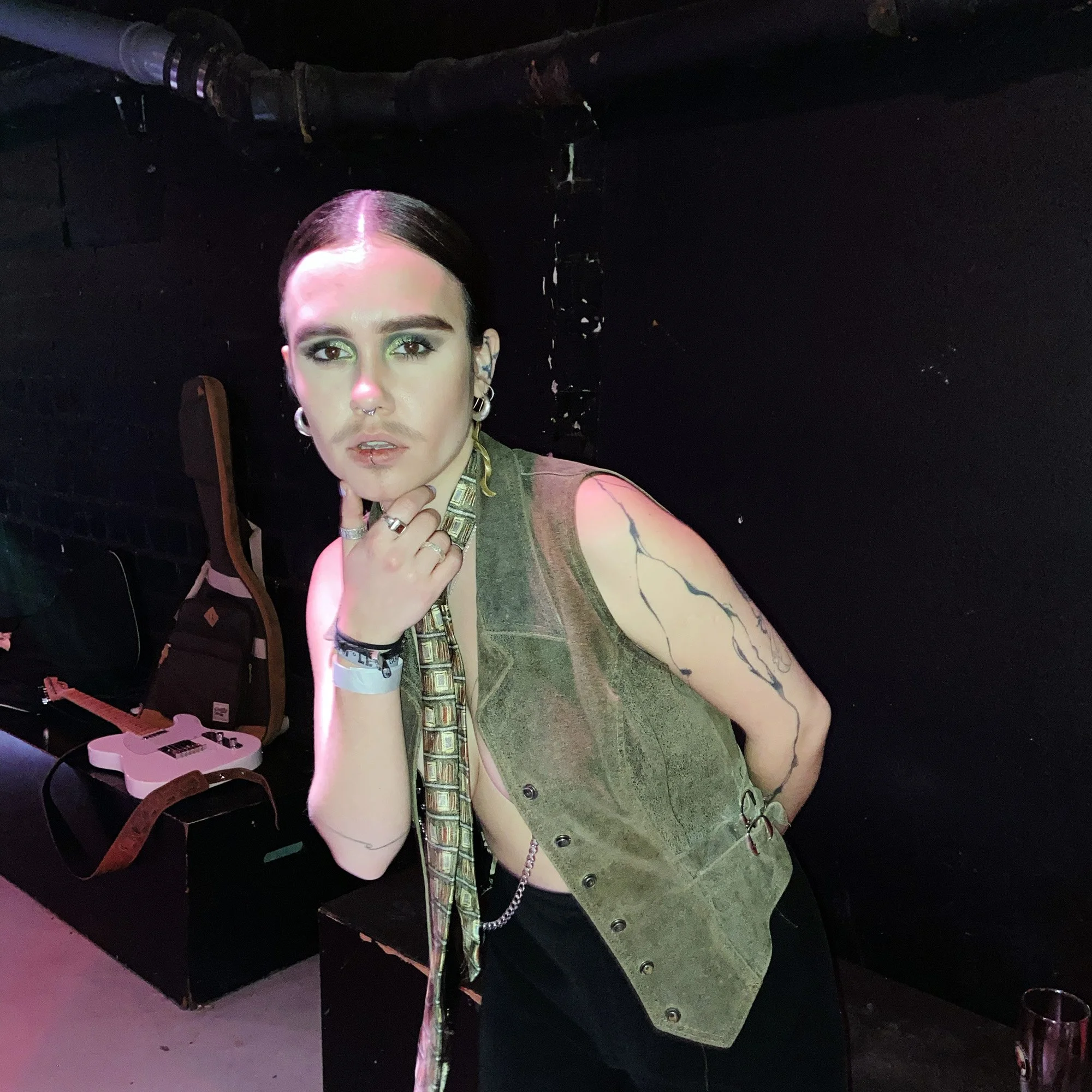 A young woman with dark hair tied back, wearing a sleeveless olive green vest, multiple rings, earrings, and a patterned scarf, poses in a dimly lit room with black walls, a guitar case and a white guitar on a table in the background, and a glass of drink on the floor.