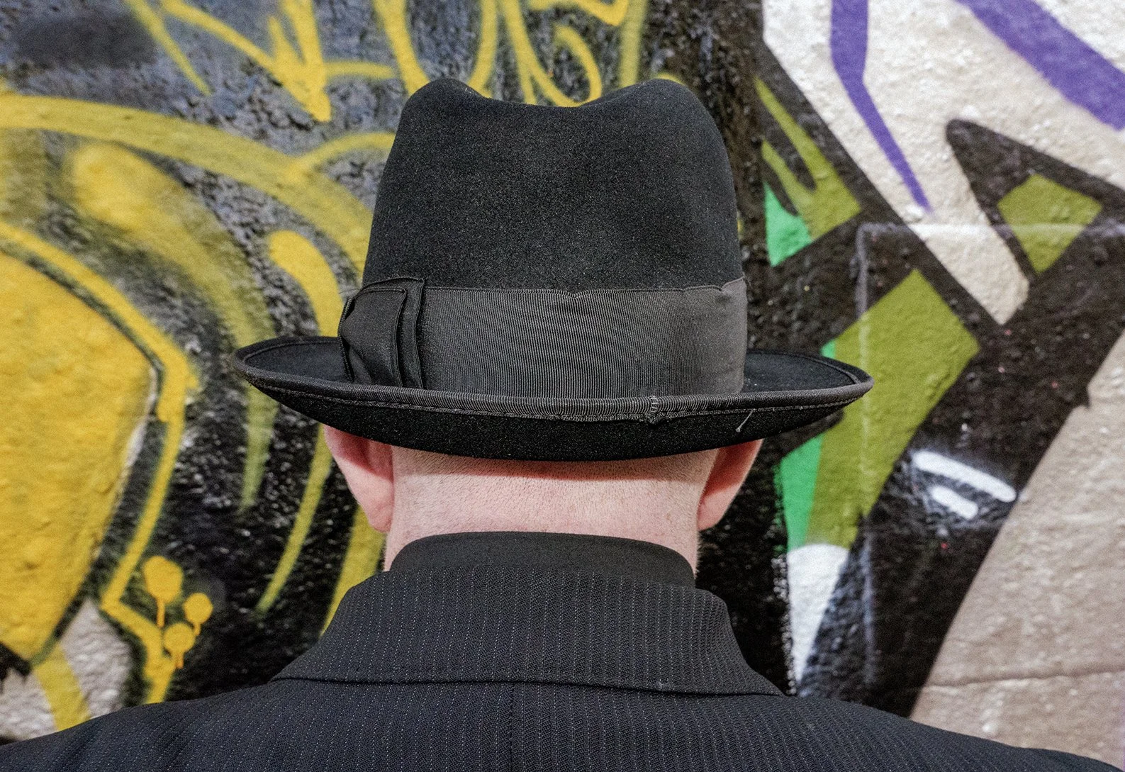 Person wearing black cowboy hat and black suit, standing in front of a colorful graffiti wall.