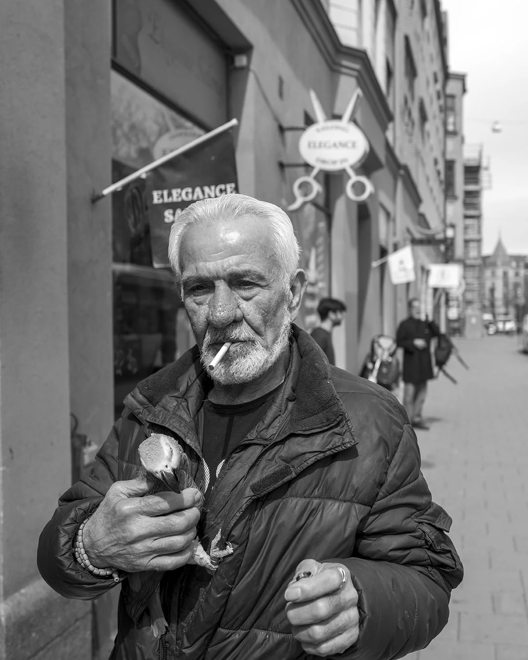 An elderly man with a beard and white hair, smoking a cigarette, holds an ice cream cone, walking on a city street in front of a shop named 'Elegance Salon.' The photo is in black and white.