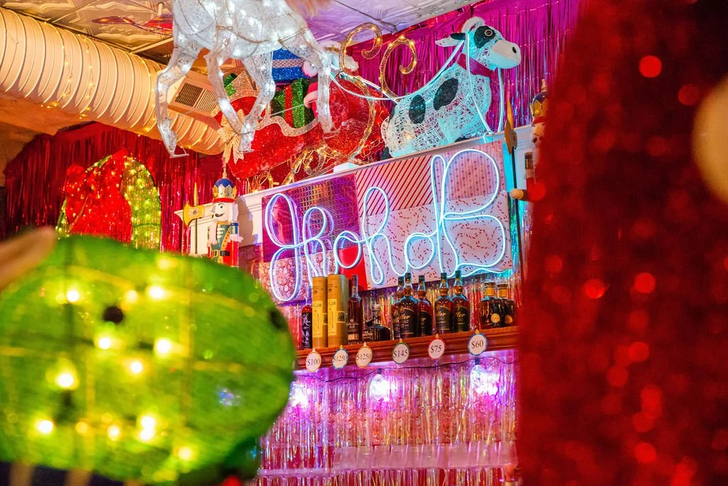 Colorful bar with Christmas decorations, including a neon sign that spells out "Cheers", a green ornament in the foreground, and a red glittery object on the right.
