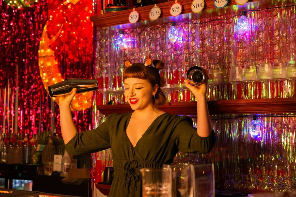 A young woman behind a bar, shaking a cocktail shaker with a smile, in a colorful, festive setting with tinsel, lights, and drink prices displayed overhead.