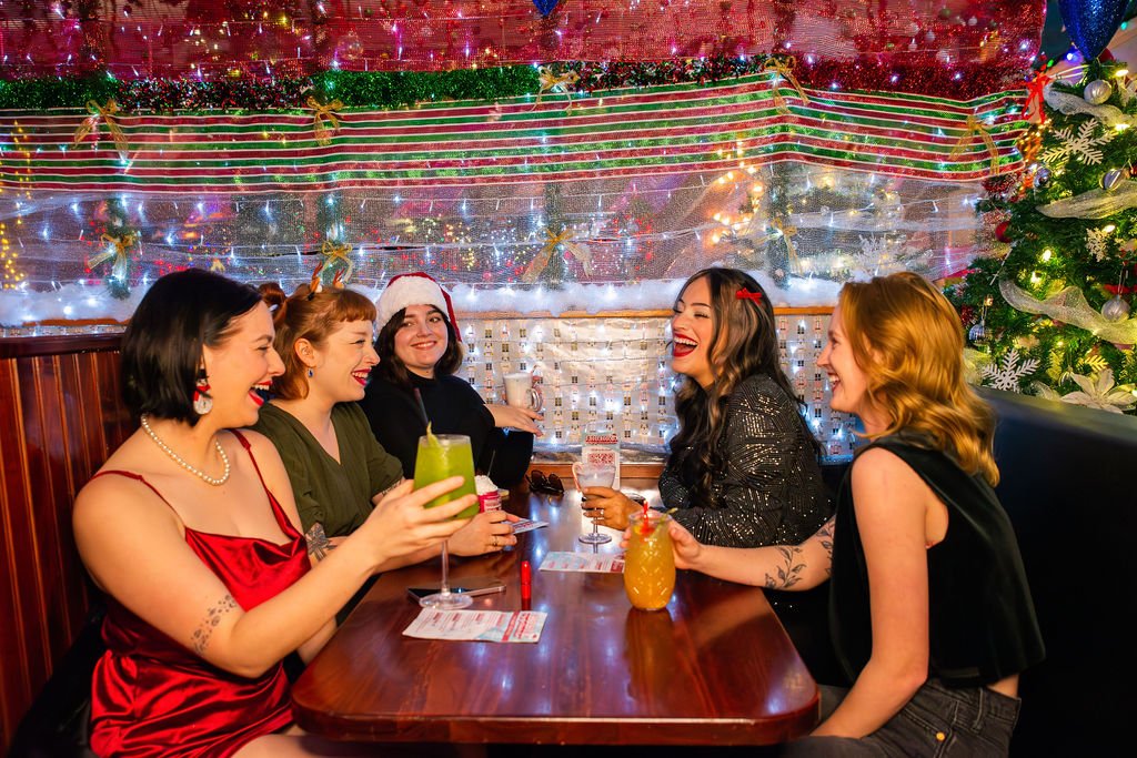 Five women sitting at a festive Christmas-themed restaurant table, laughing and enjoying drinks. One woman wears a red dress and pearl necklace, another in a green dress, a third with a Santa hat, and two others with dark hair and tattoos. The background features a decorated Christmas tree and holiday decorations.
