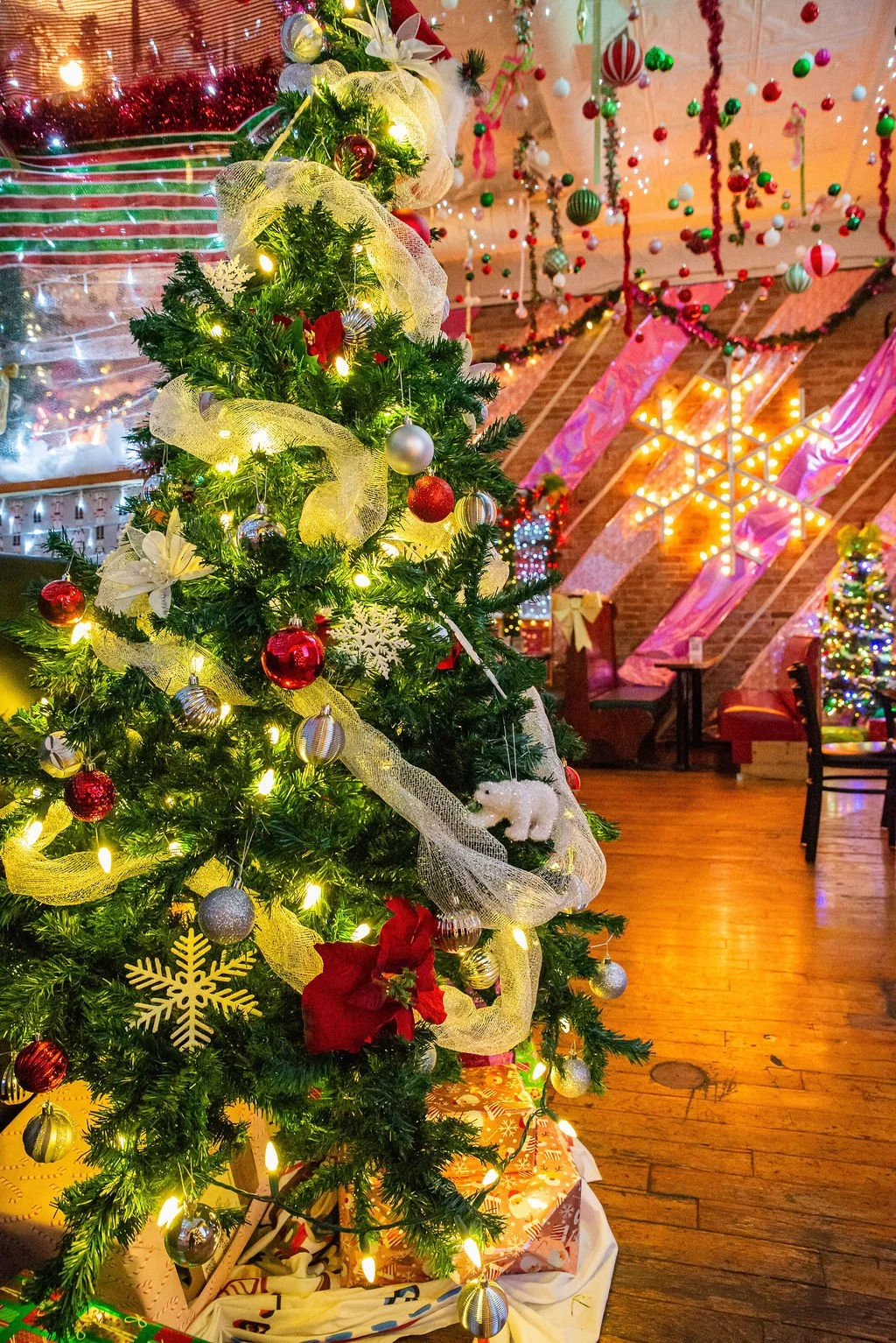 Decorated Christmas tree with ornaments and lights in a festive room with hanging decorations, a star-shaped light installation, and furniture.