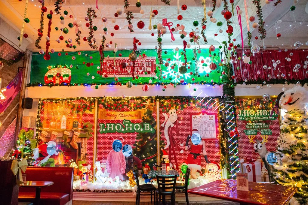Colorful Christmas decorations with hanging ornaments, garlands, and lights inside a festive restaurant. Features a Christmas scene with Santa dolls, a decorated Christmas tree, and holiday-themed banners reading "Merry Christmas" and "JHOHO B." Christmas stockings, reindeer figures, and a bright snowflake display add to the holiday ambiance.