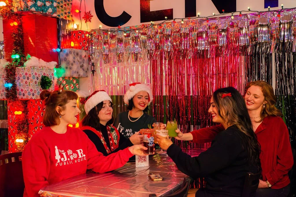 Group of five women celebrating Christmas at a party, wearing Santa hats and festive attire, raising drinks together in front of colorful holiday decorations.