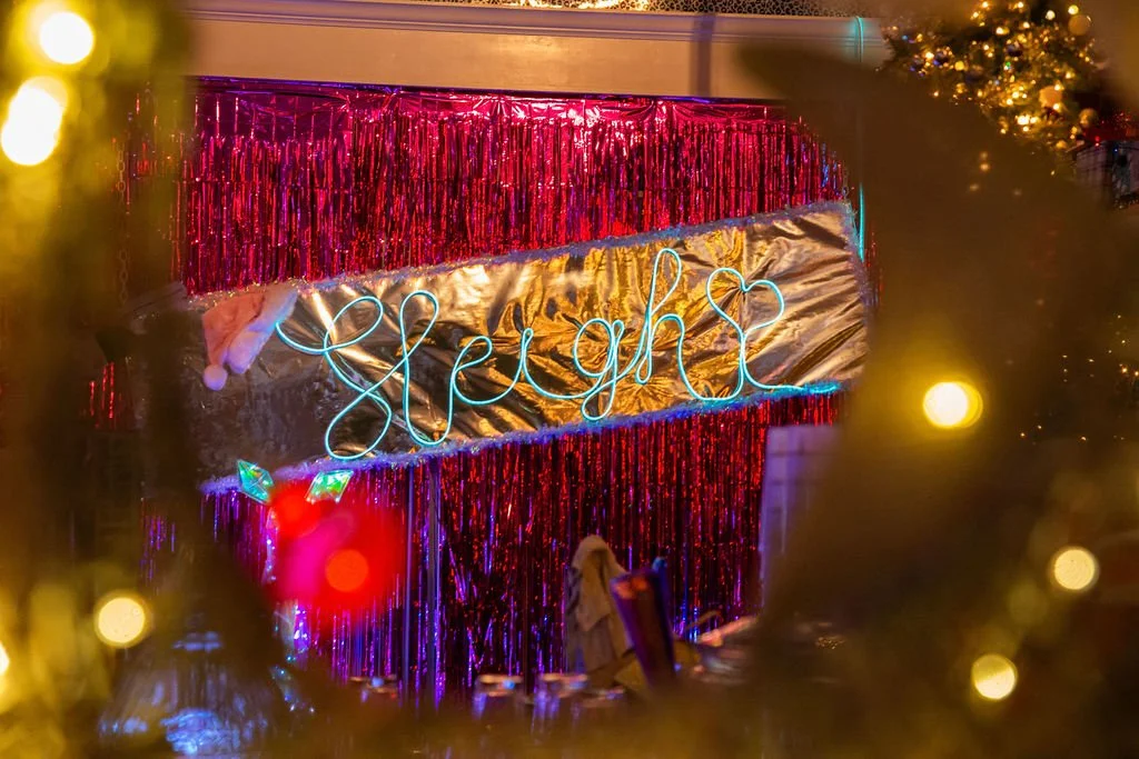 A holiday decorated space with a neon sign spelling 'sleigh' on a wall covered with metallic gold fabric and red tinsel, and Christmas lights and ornaments visible.