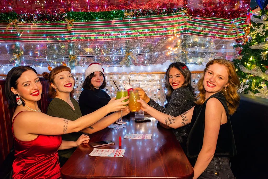 Five women celebrating Christmas at a restaurant, wearing festive clothing and drinks in hand, with Christmas decorations and a Christmas tree in the background.