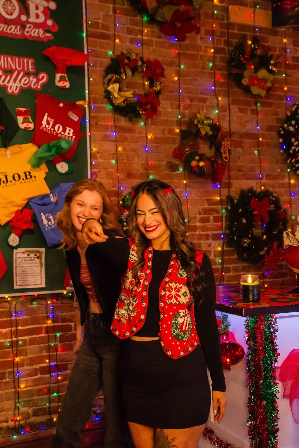 Two women smiling and laughing in a festive Christmas setting with holiday decorations, colorful lights, and gift bags in the background.