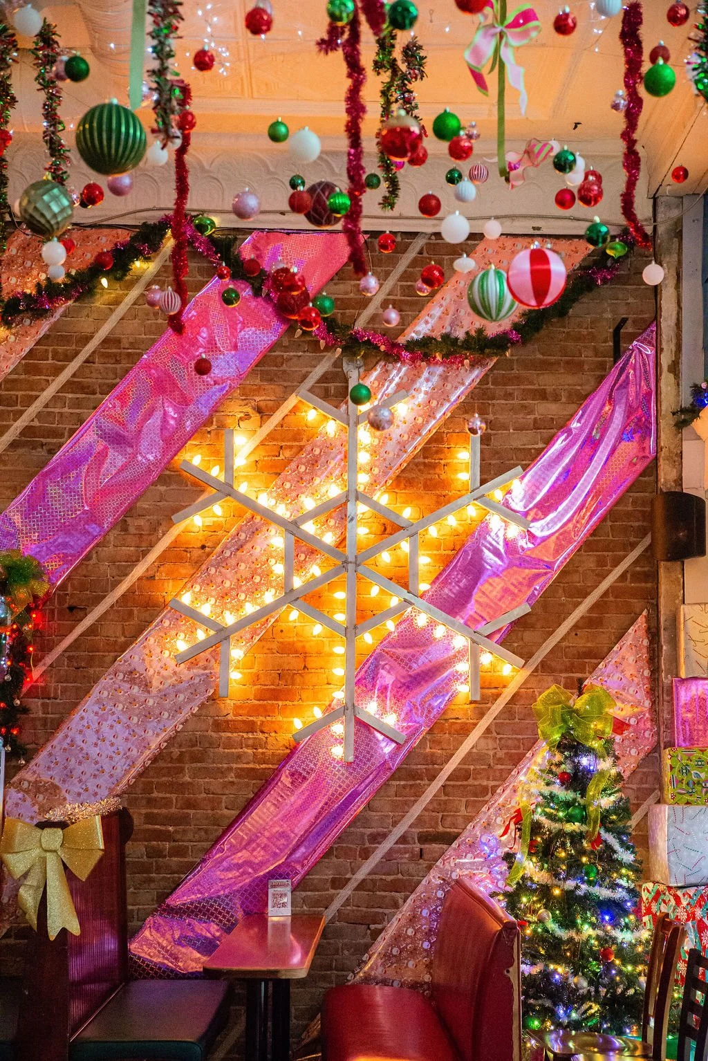 Decorated Christmas scene with hanging ornaments, tinsel, a Christmas tree with lights, presents, and a large illuminated star on a brick wall.