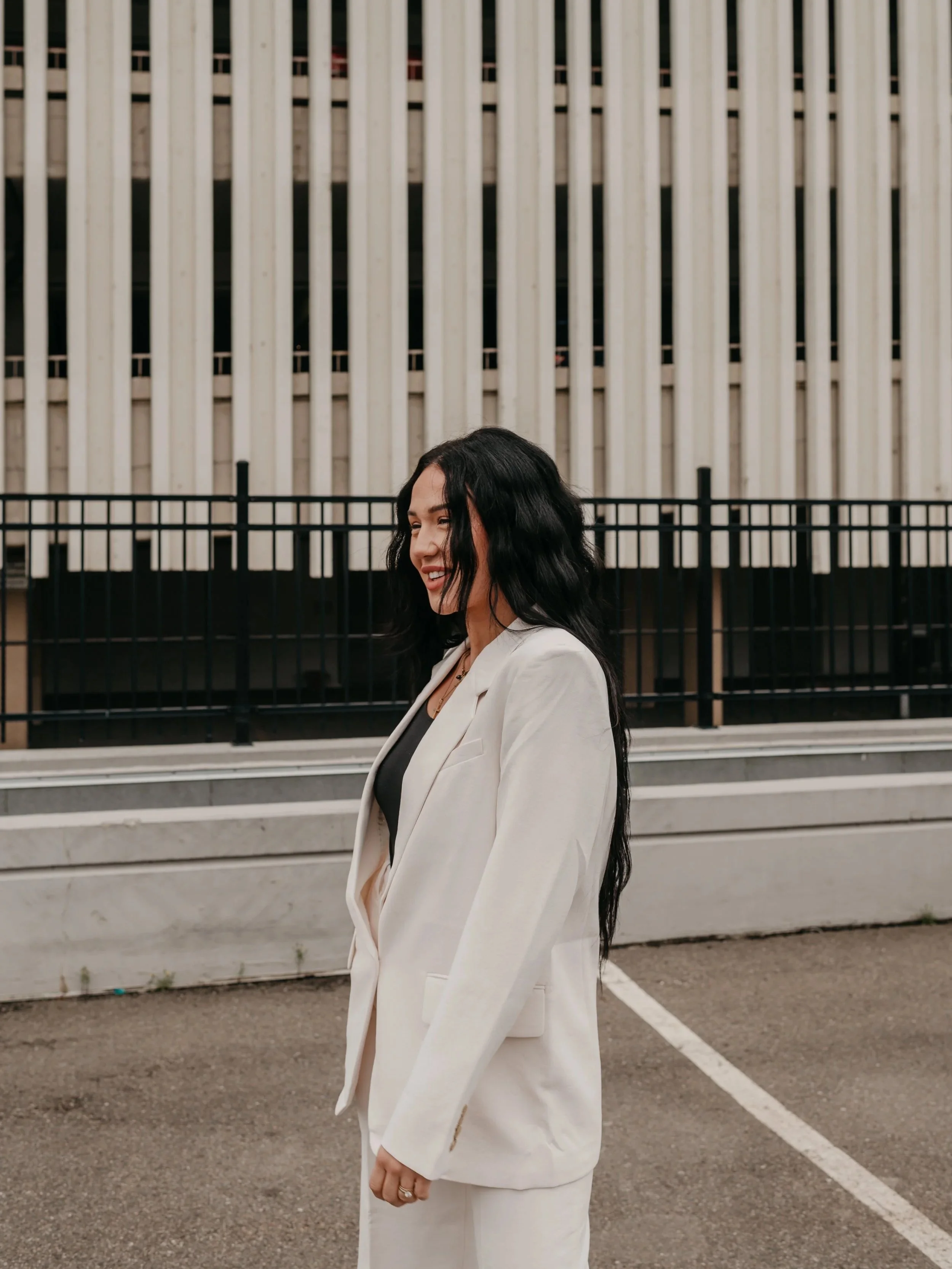 A woman with long black wavy hair smiling and looking to the side, standing outdoors in front of a modern building with vertical white architectural elements, wearing a white suit jacket over a black top.