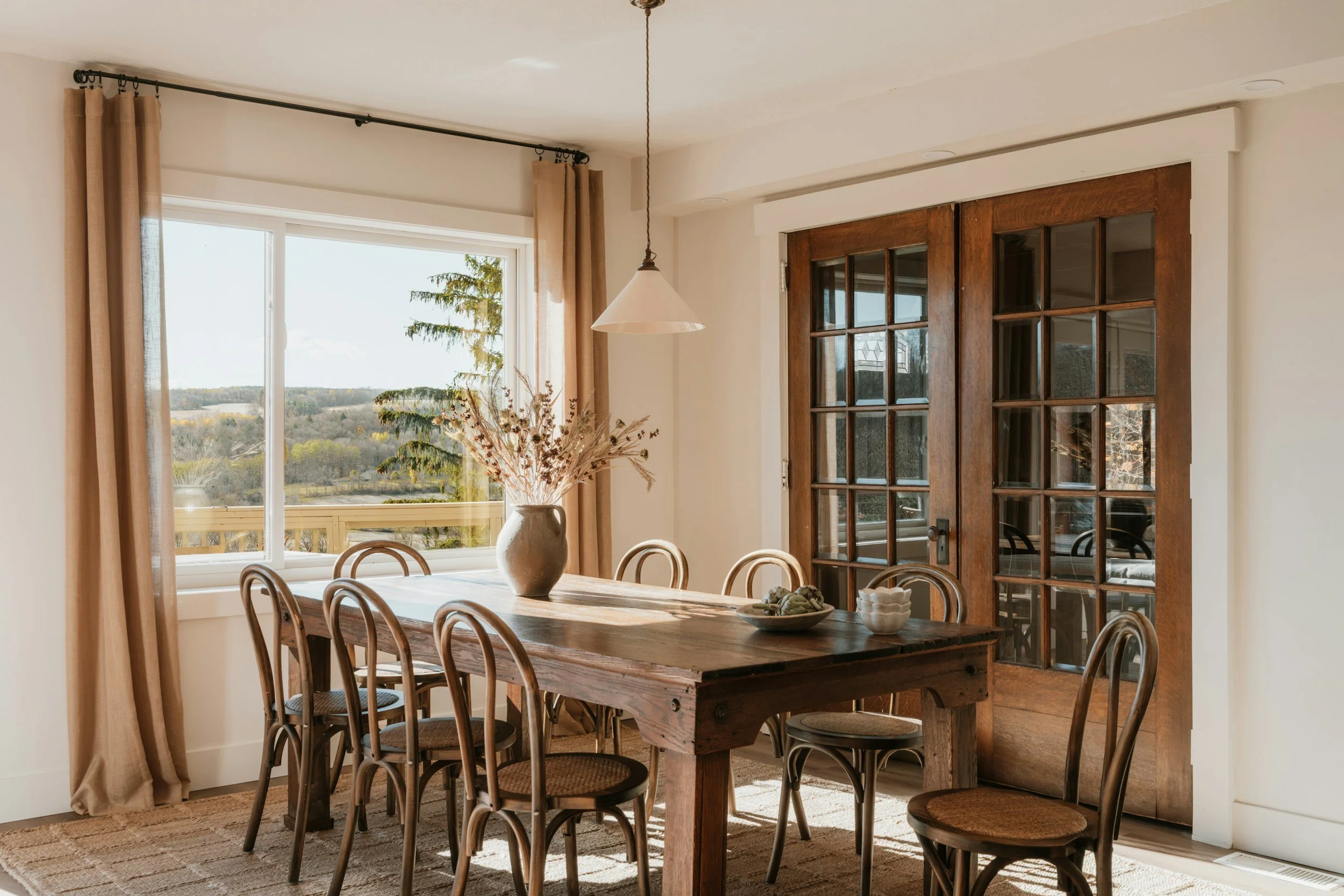 A dining room with a wooden table, six matching chairs, a large vase with dried flowers, a bowl, and a small dish on the table. A large window with beige curtains shows a landscape outside, and there are glass-paneled wooden double doors next to the window. The room is well-lit with natural light.
