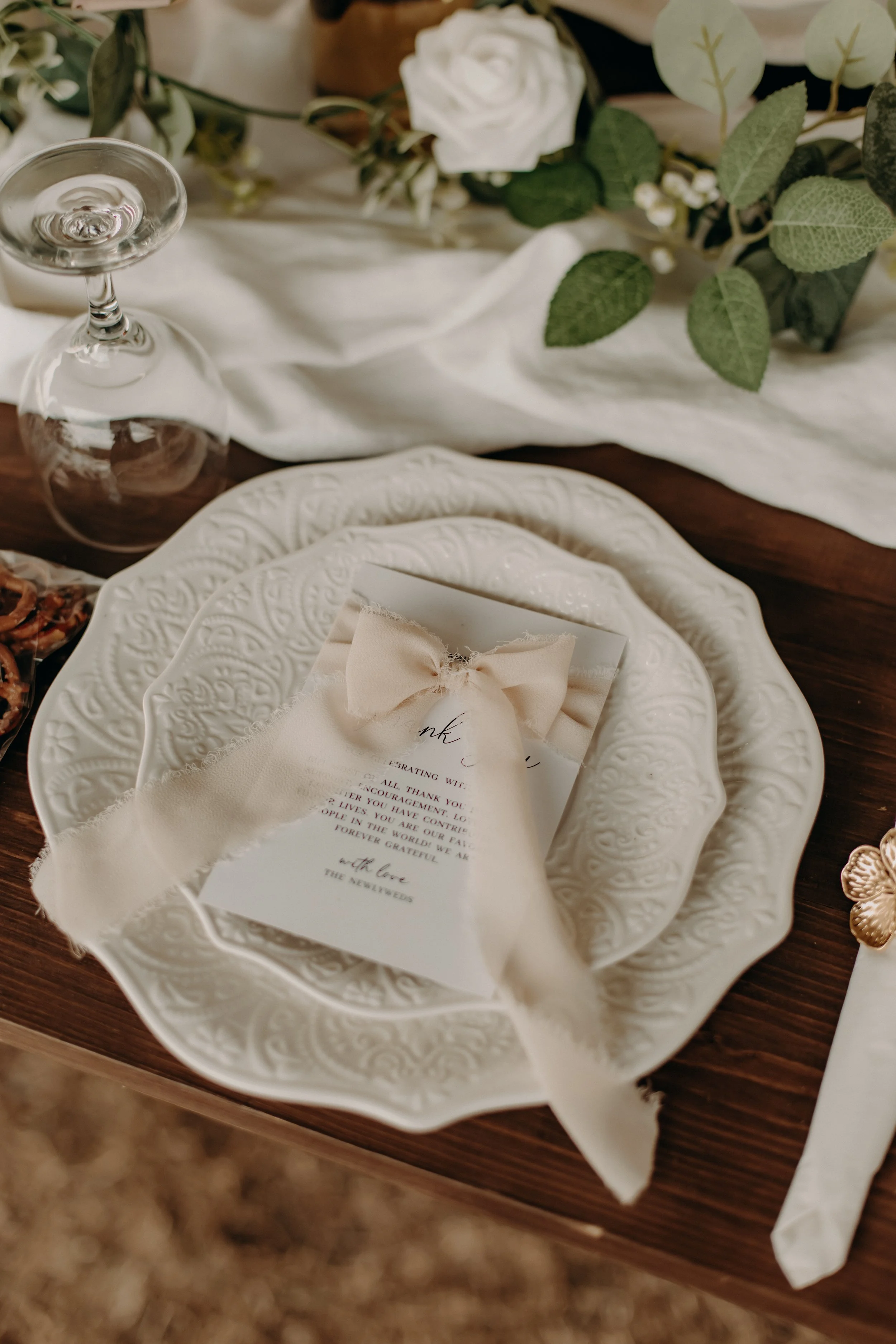 Elegant table setting with a white decorative plate, a cream-colored napkin with a bow, a menu, a glass turned upside down, and floral decorations with white roses and green leaves.
