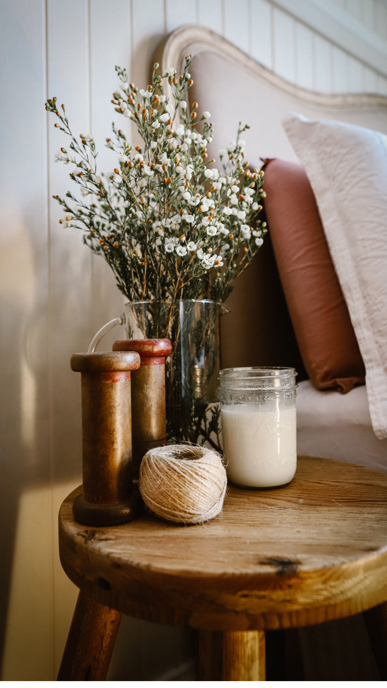 Decorative wooden side table with candles, twine, and a vase of white flowers in a cozy, softly lit bedroom.