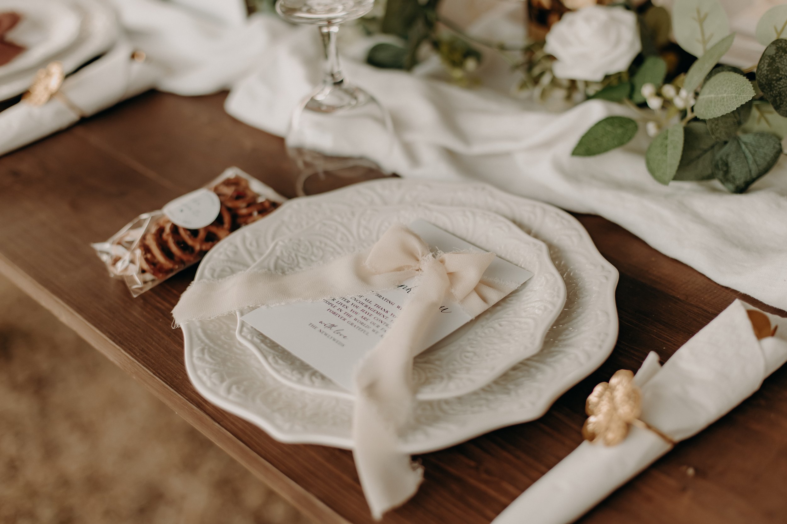 Elegant table setting with ornate white plates, a menu card tied with a cream ribbon, a wrapped napkin, and decorative greenery on a wooden table.