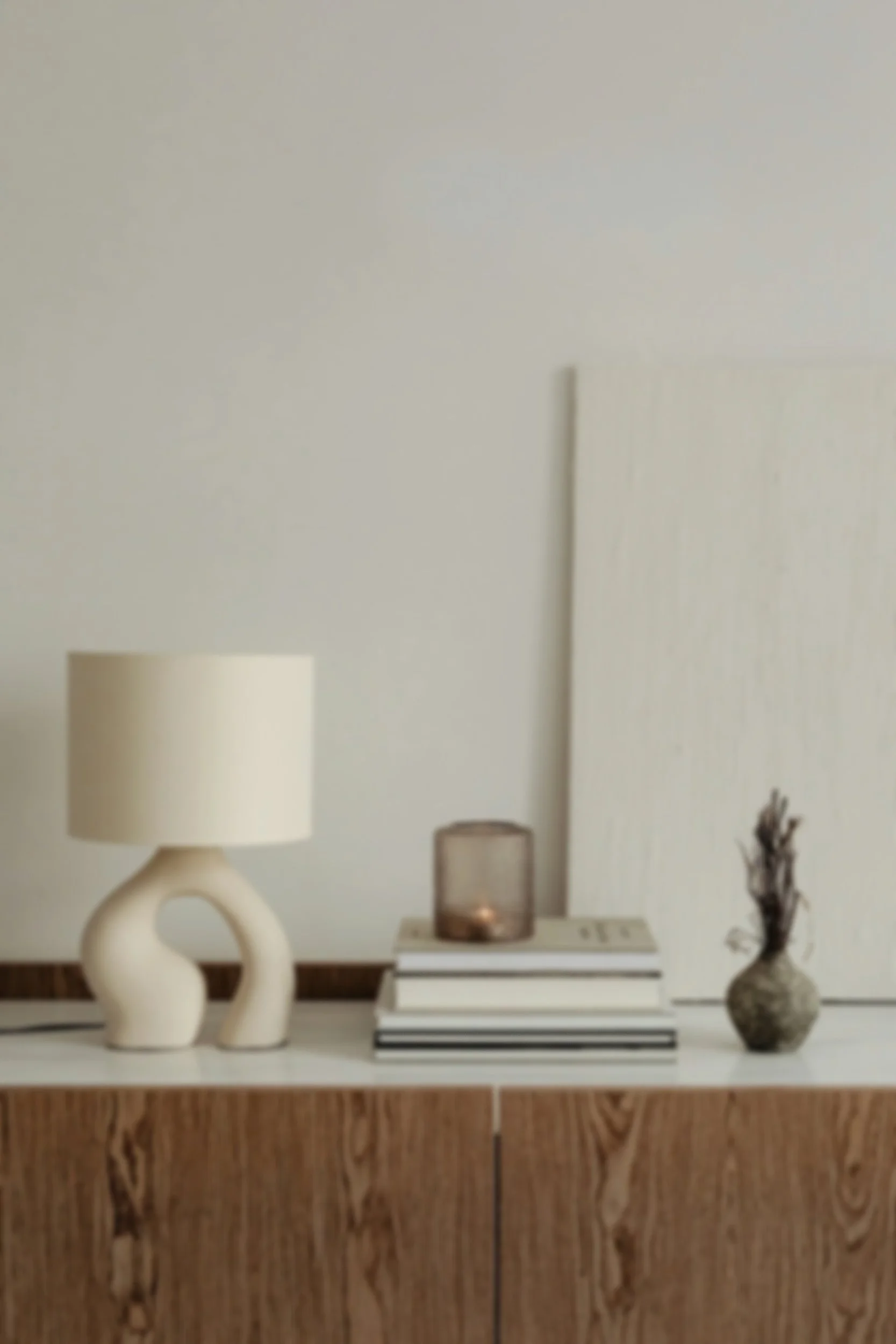 A minimalist sideboard with a cream-colored sculptural table lamp, a small candle in a brown glass holder, a stack of books, and a small decorative vase with dried branches, set against a plain beige wall.