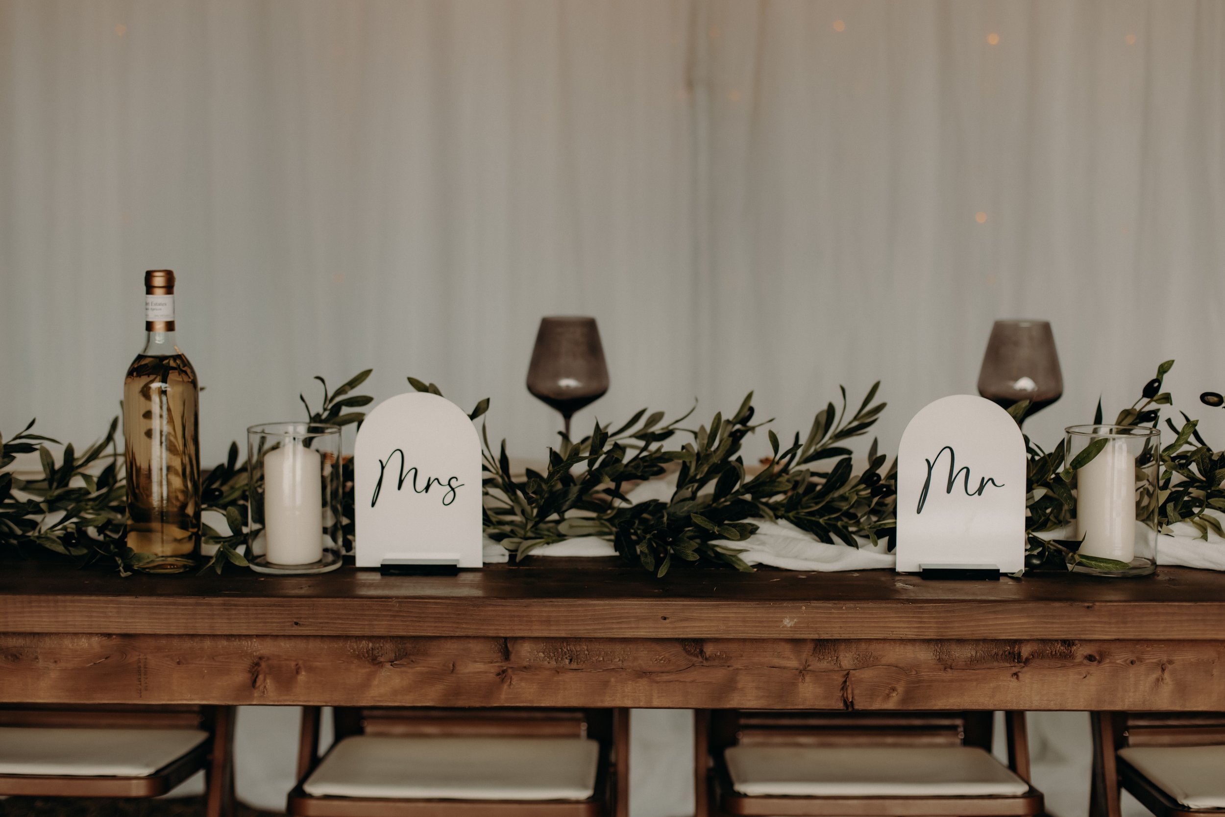 Wedding reception table with a wooden surface, greenery garland, candles, wine glasses, a bottle of alcohol, and signs labeled 'Mrs' and 'Mr'.