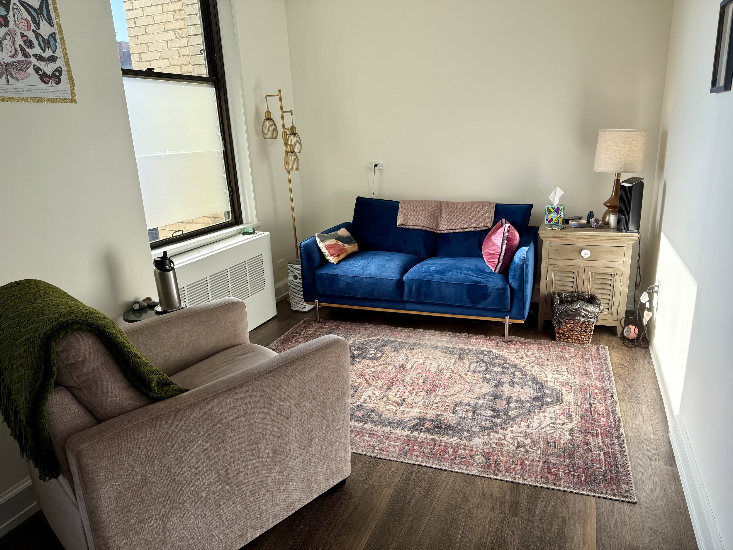 A living room with a beige couch with a green blanket, a small dark table, a window, a blue velvet sofa with pillows and a throw, a wooden side table with a lamp, decorative items, and a patterned area rug.