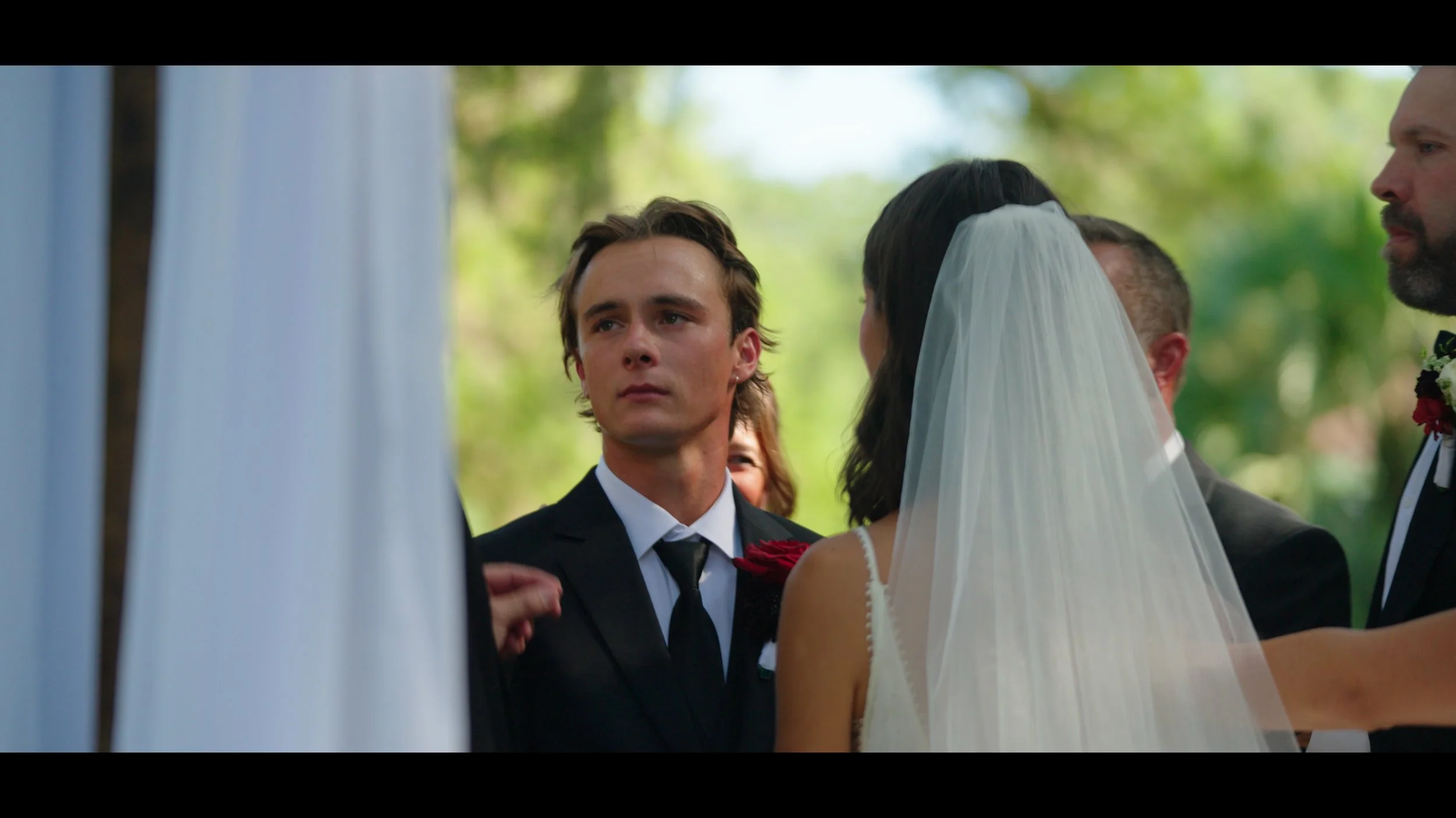 A wedding ceremony outdoors with a groom and bride, surrounded by guests. The groom has a serious expression and is dressed in a black suit with a black tie. The bride is wearing a white wedding dress with a veil.