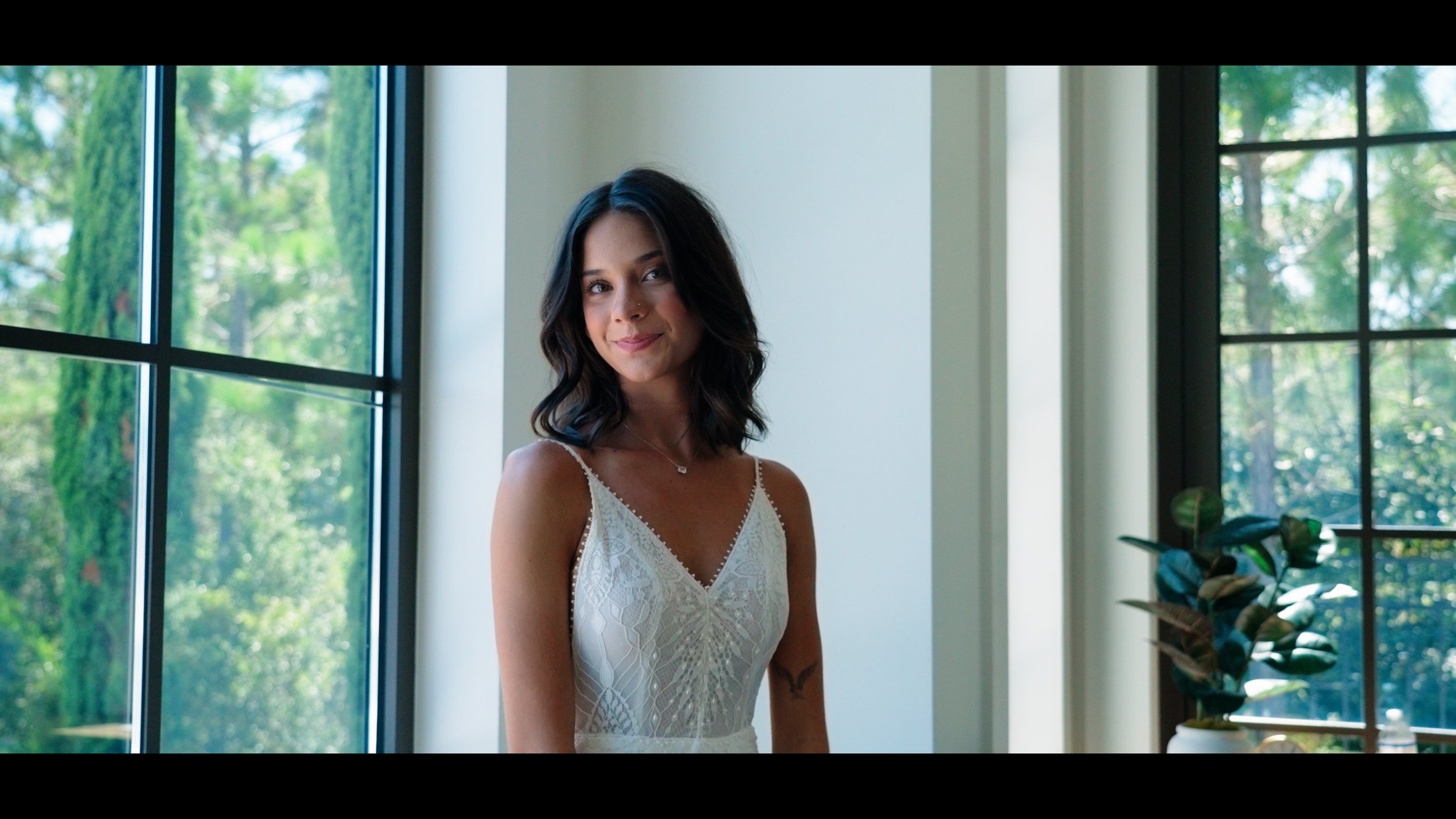 A woman with shoulder-length dark hair wearing a white dress with lace detailing, standing indoors near a window with greenery outside, smiling softly at the camera.