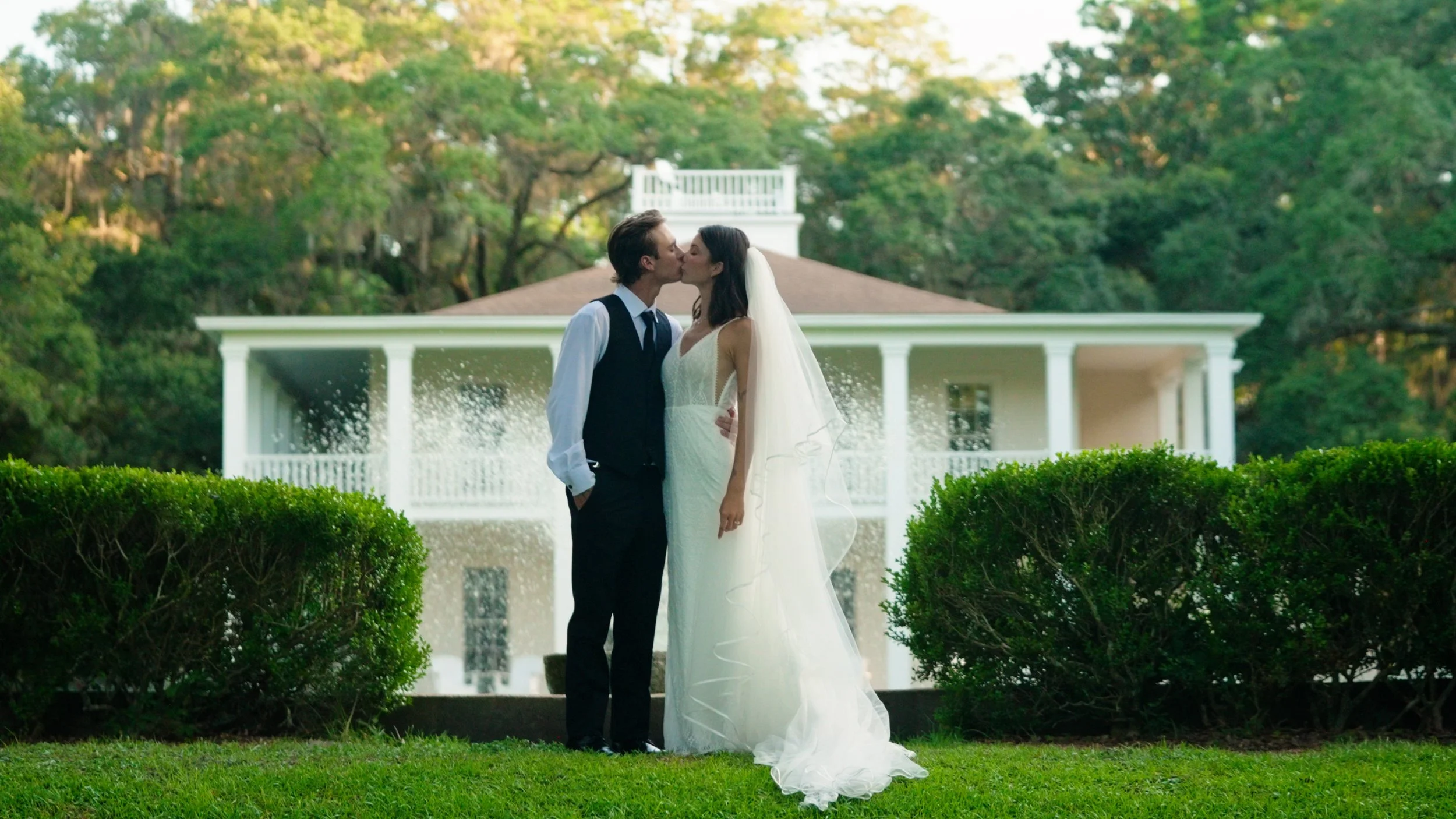 A bride and groom sharing a kiss on a lush green lawn in front of a white house with a porch. The bride is wearing a white wedding dress and veil, and the groom is in a black vest and slacks with a white shirt.