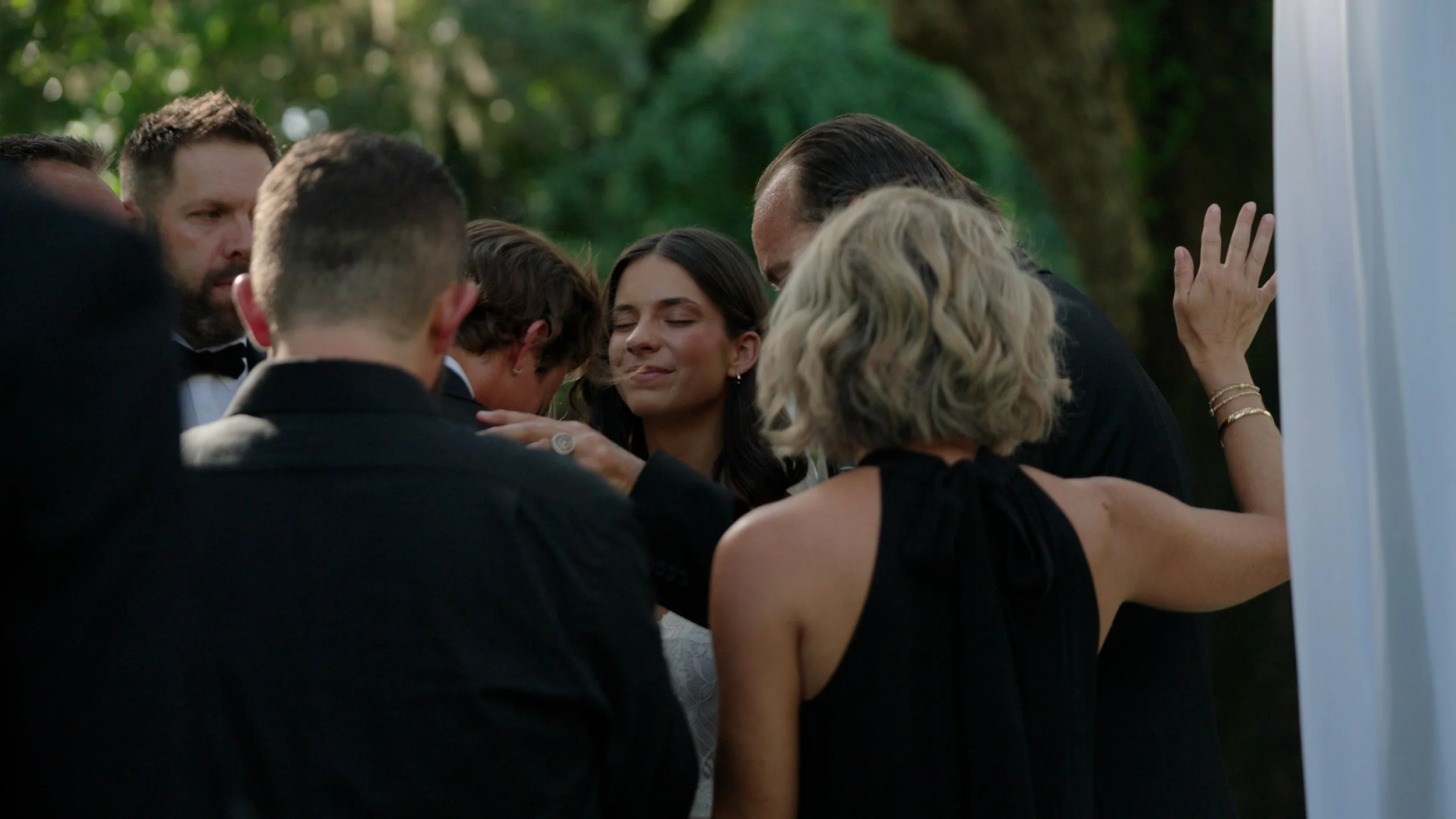 People gathered outdoors in a group hug, dressed in formal attire, with trees in the background.