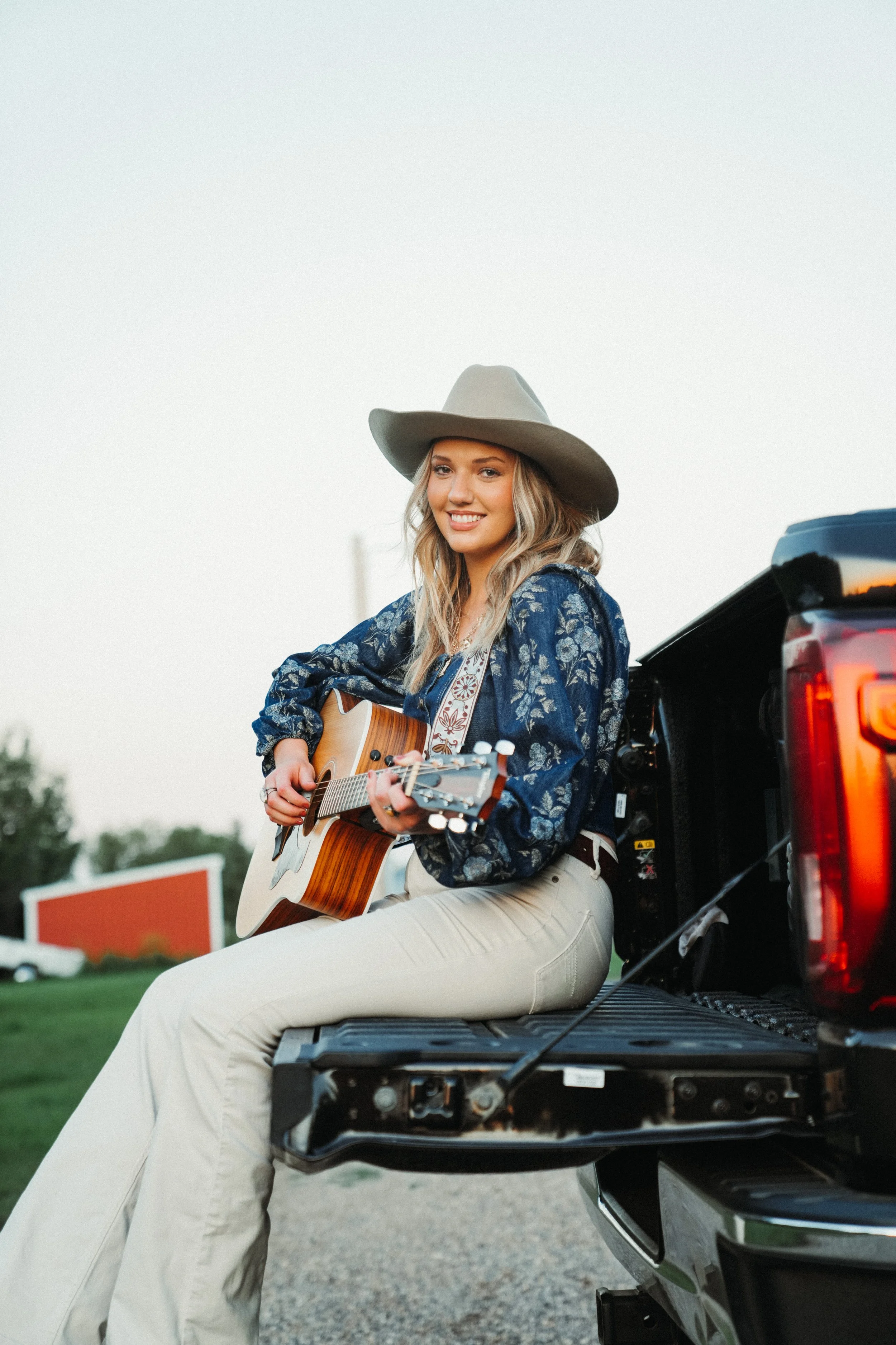 A young woman wearing a cowboy hat and a floral blouse, sitting on the tailgate of a truck, playing an acoustic guitar outdoors during sunset.